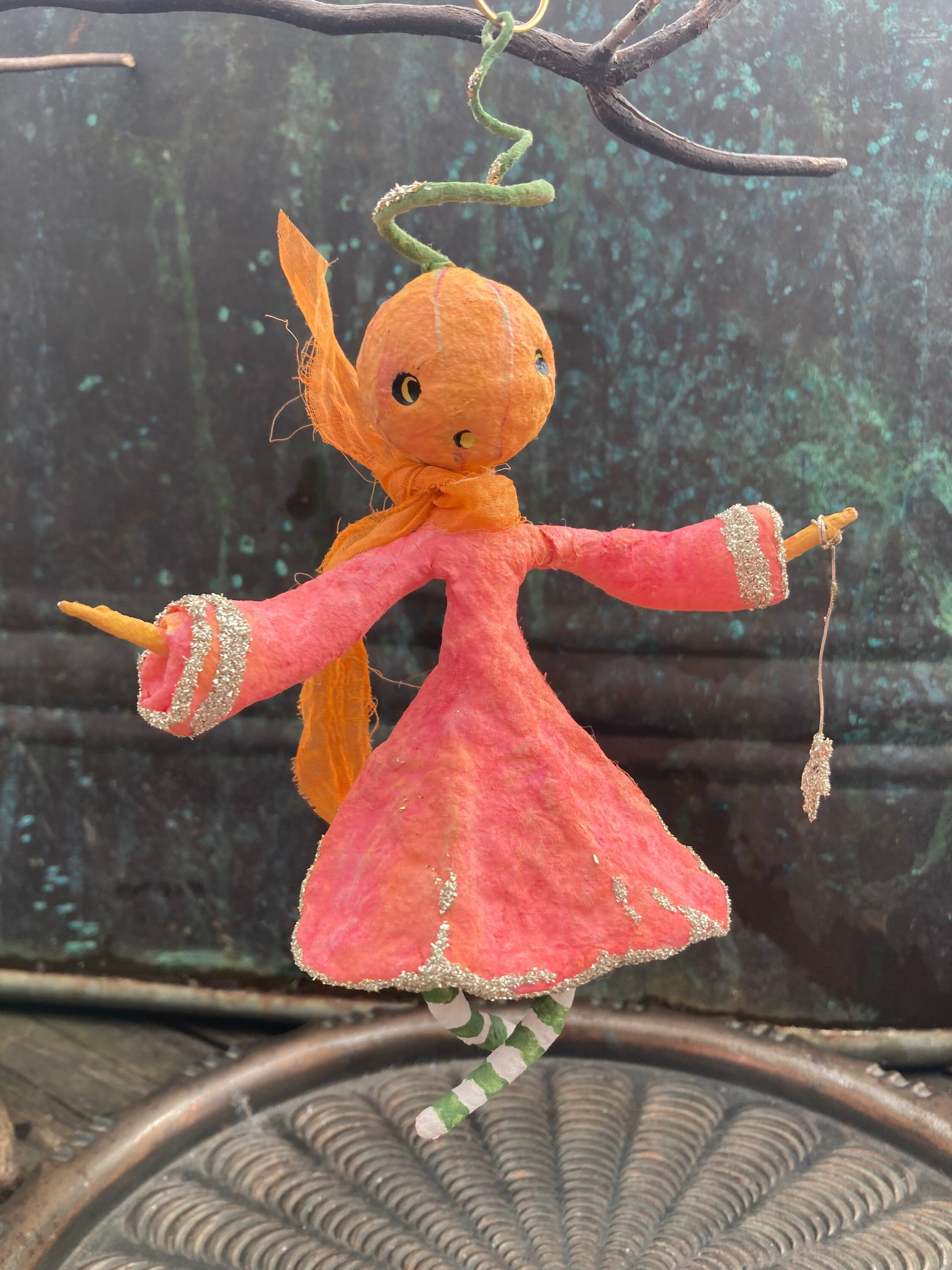 A decorative hanging doll resembling a person with a large, round orange head and shiny black eyes, wearing an orange dress with silver accents and a long striped tail, hanging from a tree branch. The background is a blurry green forest scene.