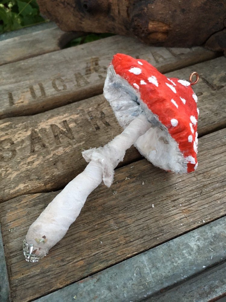 Handmade mushroom ornament with red and white cap, and white stem, placed on a rustic wooden surface.
