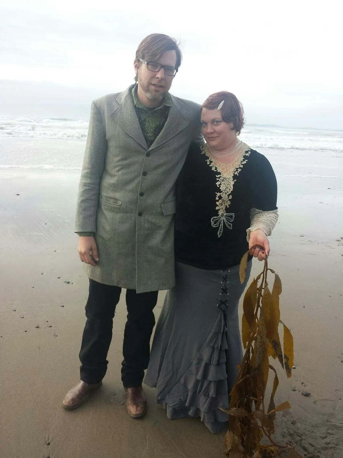 A man and a woman standing on a beach, with ocean waves in the background. The man is wearing a gray coat and glasses, and the woman is dressed in a black and gray gown with lace details. She is holding a large seaweed. The sky is overcast.