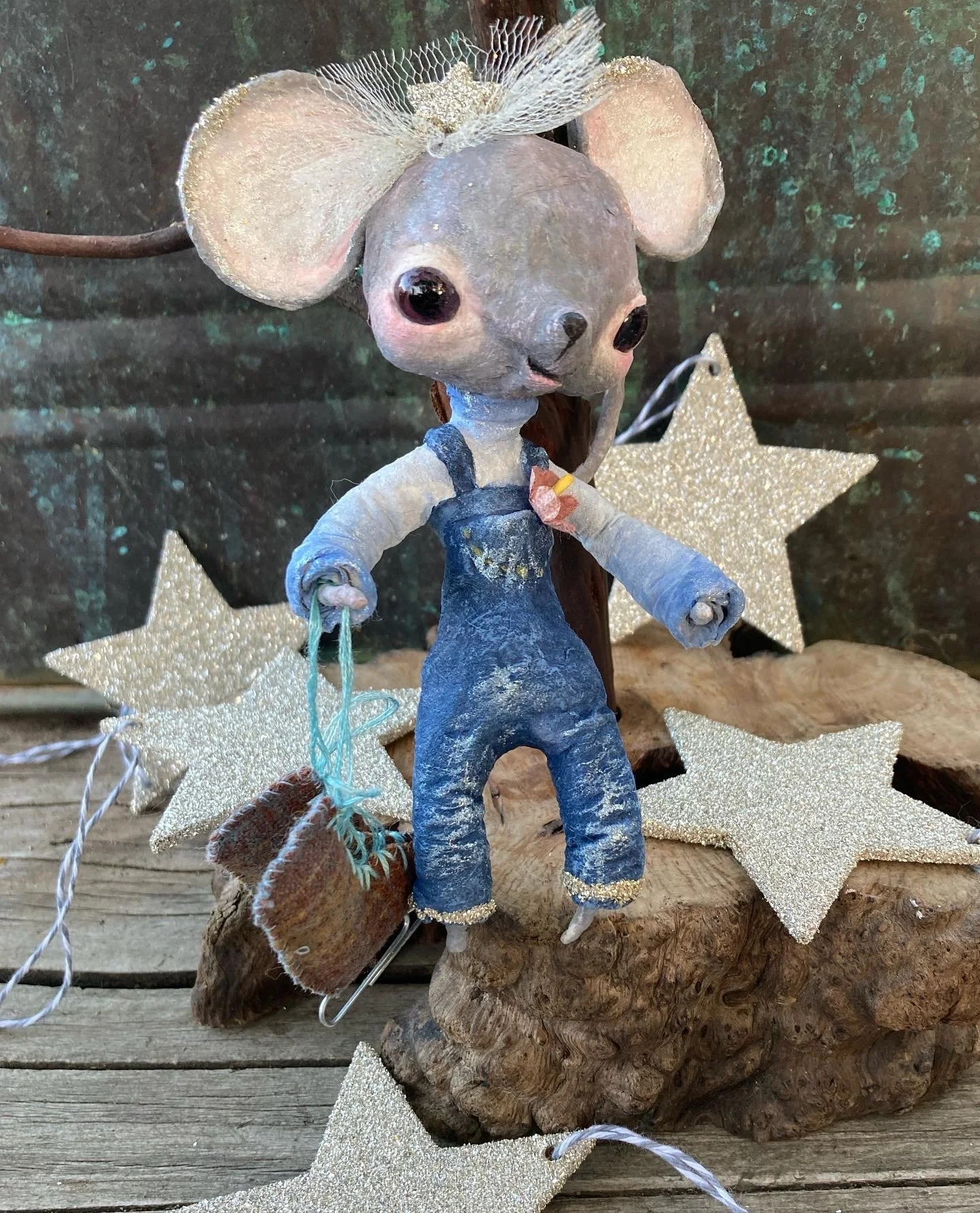 A small mouse figurine wearing blue overalls and a shirt, with large ears and shiny black eyes, standing on a piece of wood, surrounded by glittery star decorations, with a gold glittery star behind it.