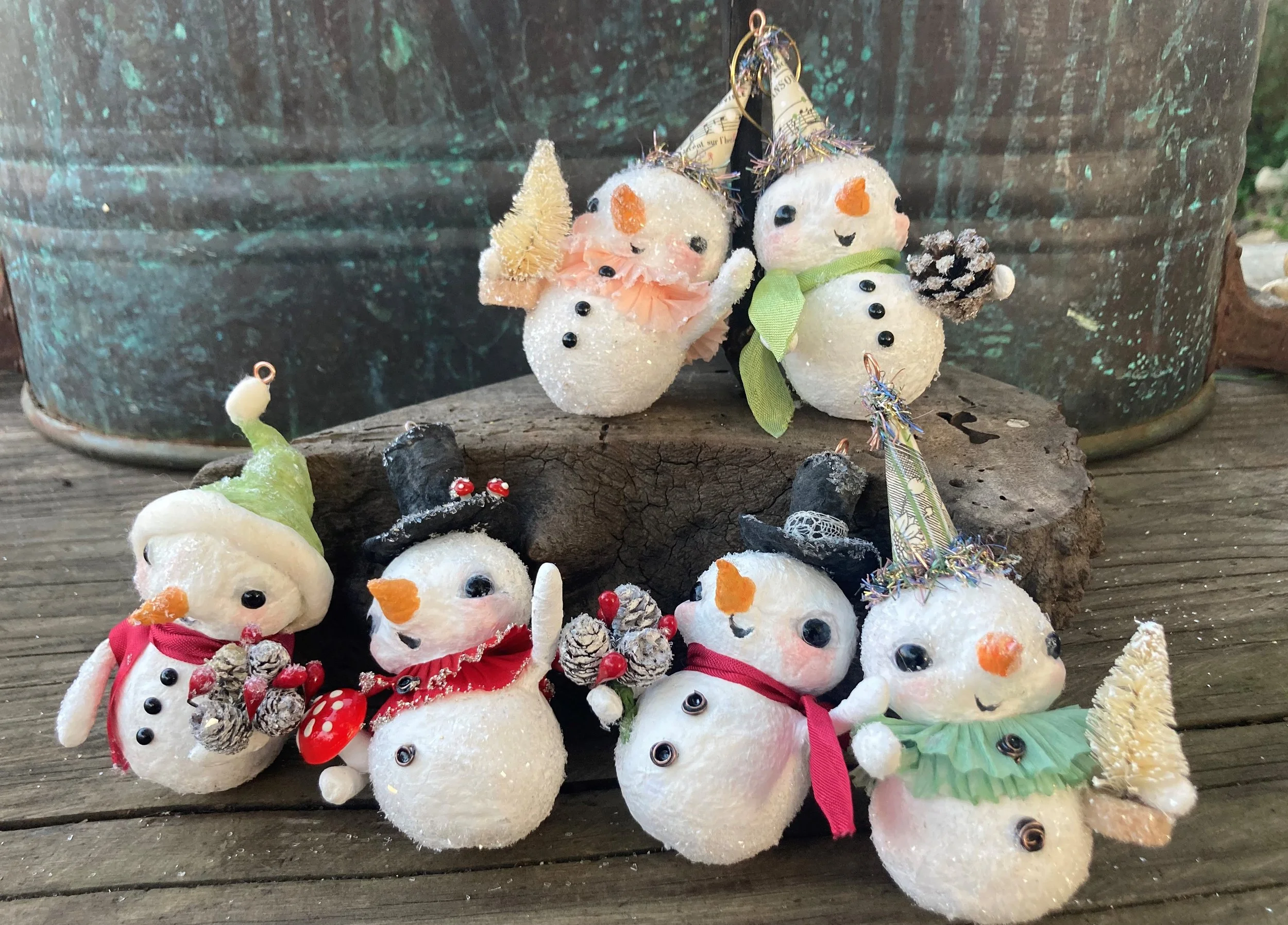 A collection of small snowman ornaments made from white fuzzy and foam materials, decorated with various accessories like hats, scarves, and pinecones, arranged outdoors on a wooden surface.