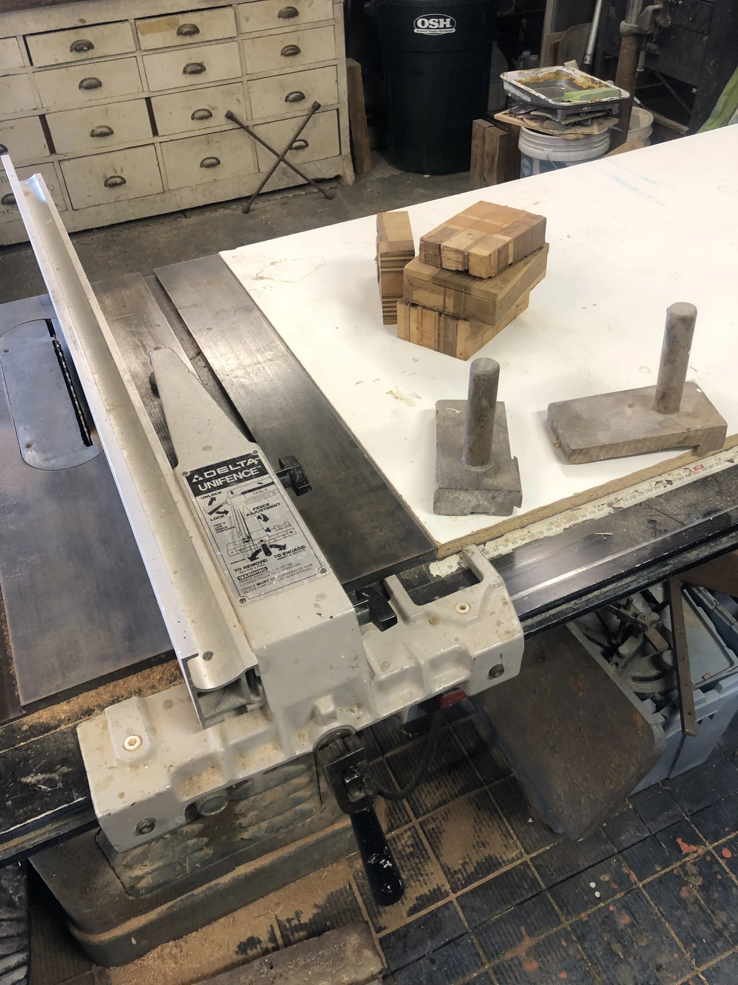 A woodworking shop with a band saw in the foreground and a stack of wooden blocks on it. There are various tools, containers, and a chest of drawers in the background.