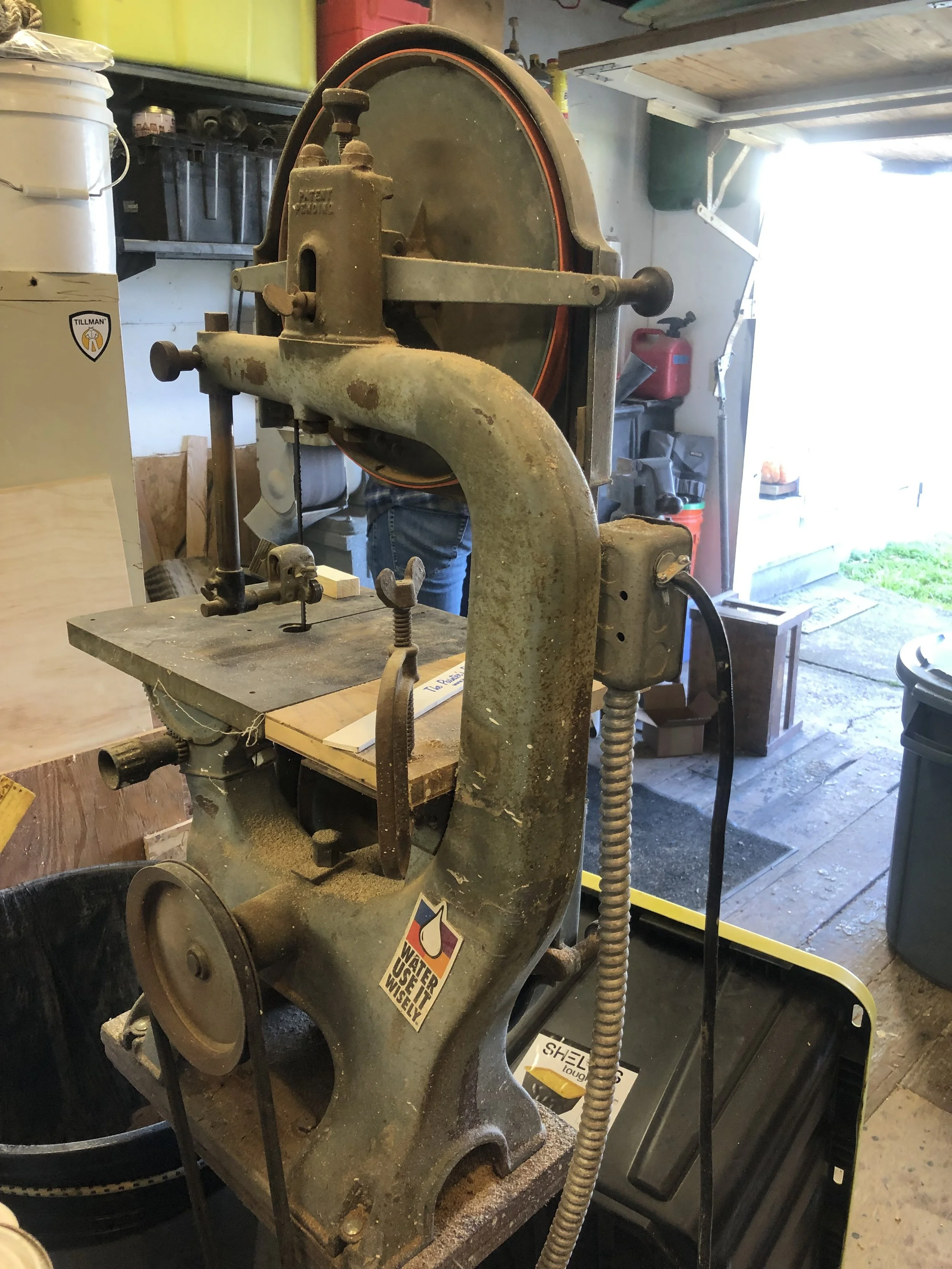 A vintage bandsaw in a workshop with various tools and supplies on shelves in the background.