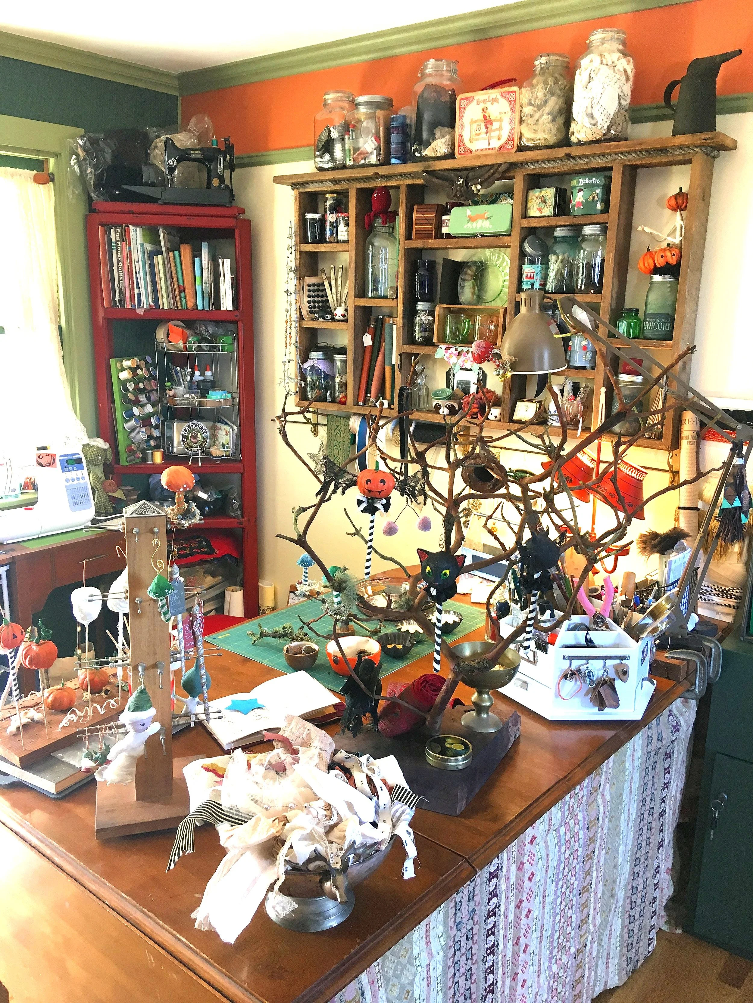 A cluttered craft or work table filled with handmade decorations, small figurines, jars, and various craft supplies. In the background, there are wall shelves filled with jars, books, and decorative items. The room has green and orange walls with a p