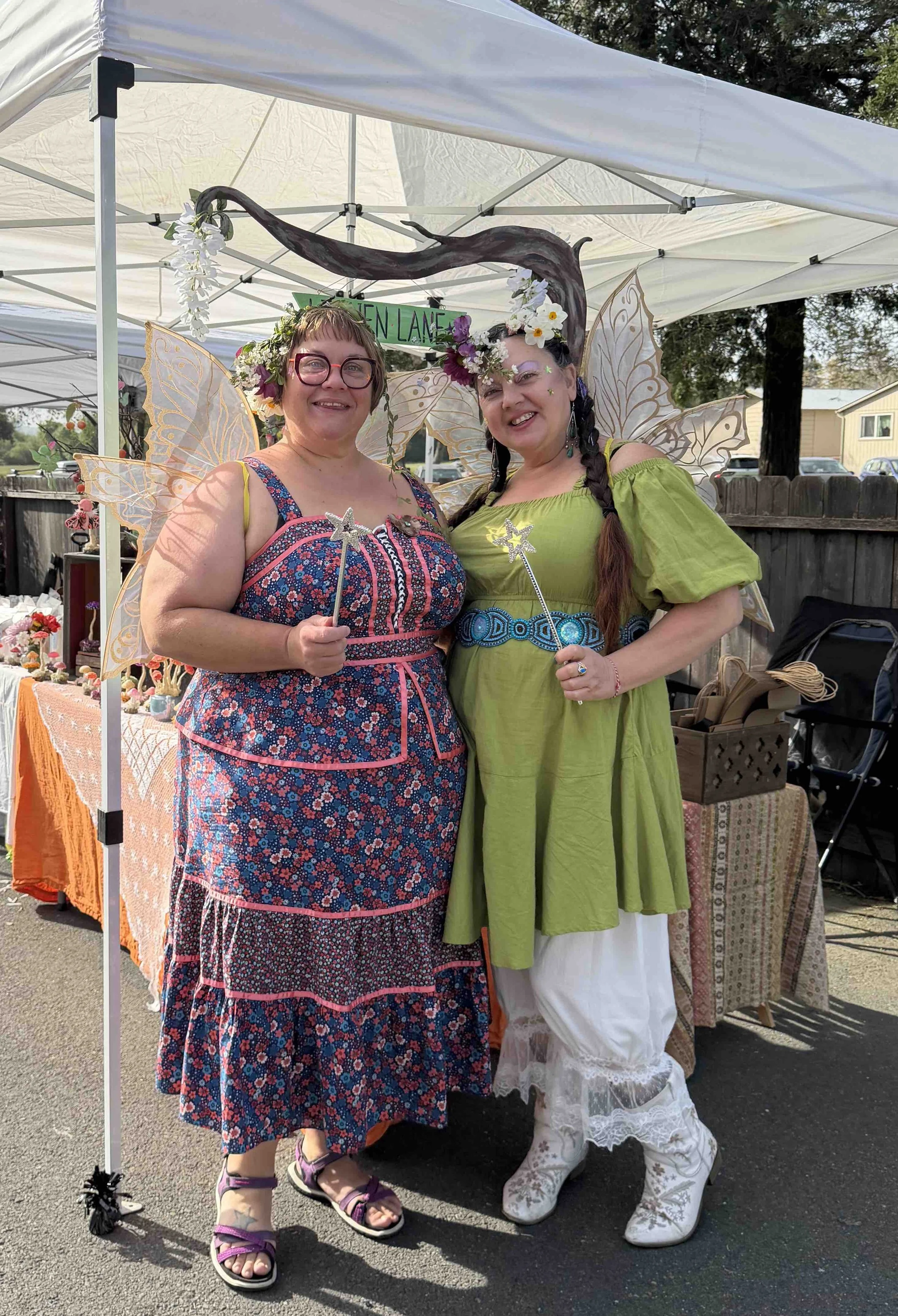 Two women dressed in fairy costumes, one with butterfly wings and the other with fairy wings, are smiling and holding magic wands at an outdoor event.