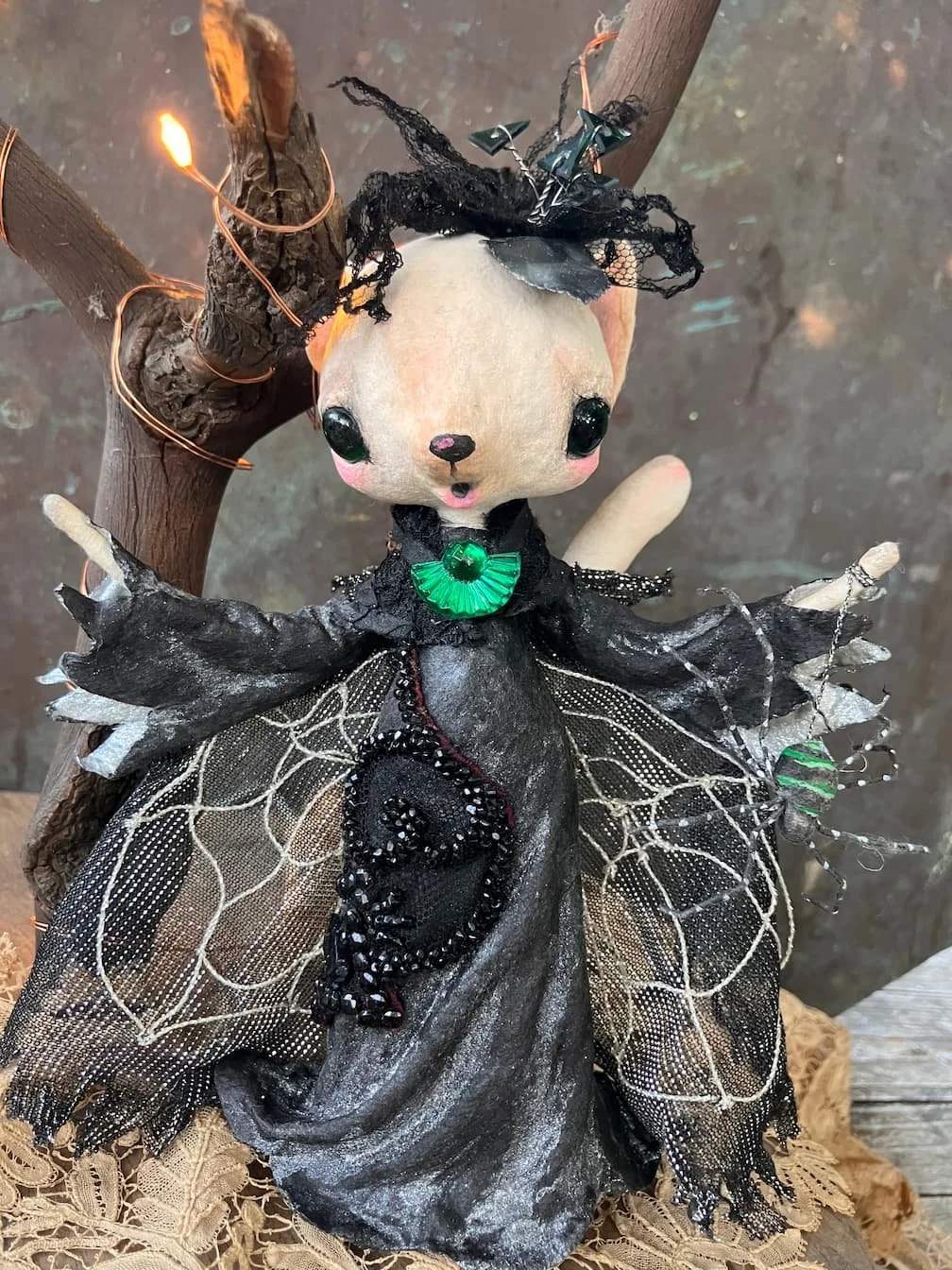 A spun cotton ermine ornament dressed as a witch with black spiderweb wings, a black lace dress, green jewelry, and black lace fabric, positioned in front of a small tree with string lights.