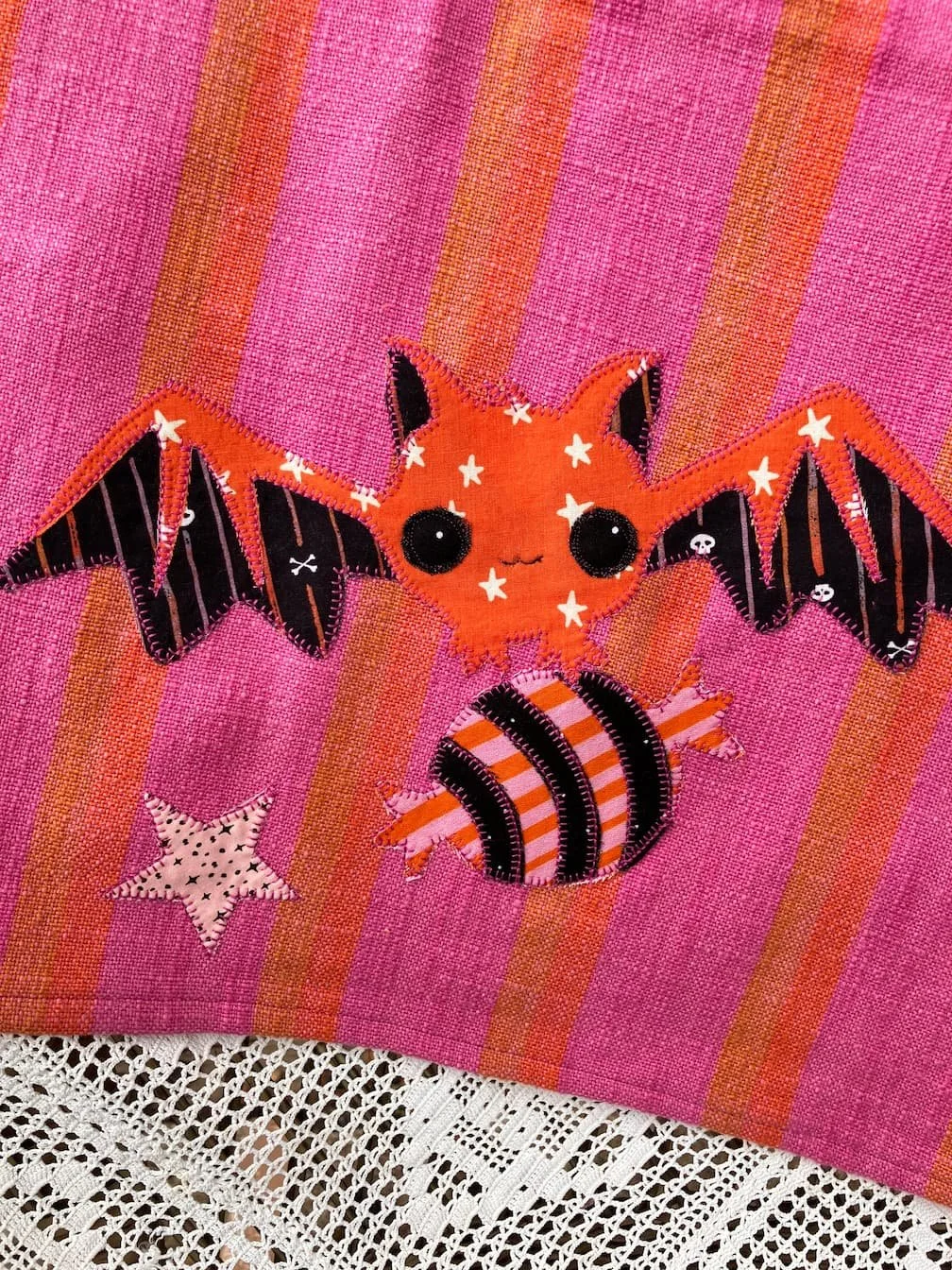 A pink fabric tea towel with orange and black embroidered bat, star, and striped raccoon patches.
