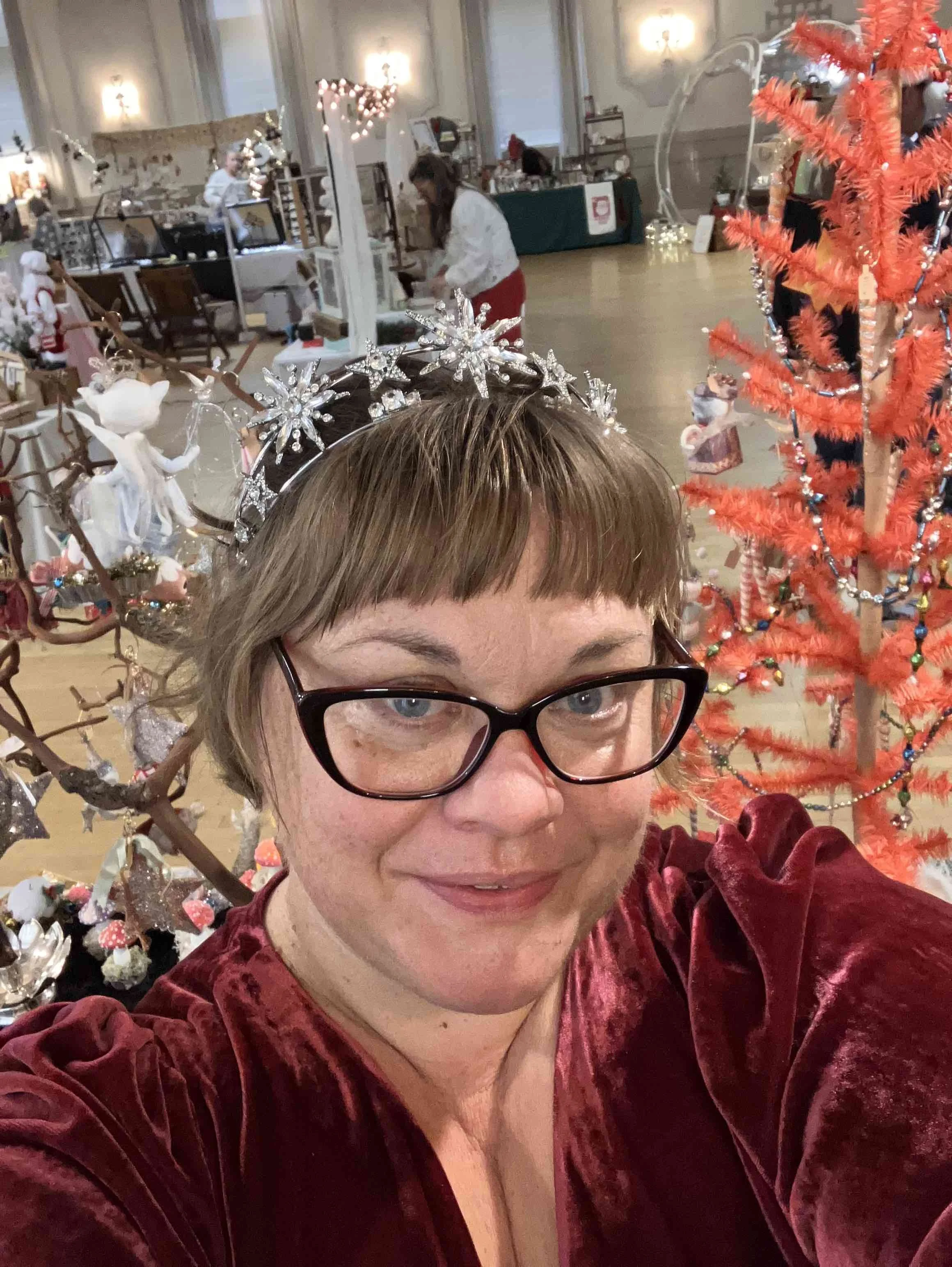 A woman with glasses and short brown hair in a red velvet dress taking a selfie at a Christmas event, with Christmas decorations and a decorated tree in the background.