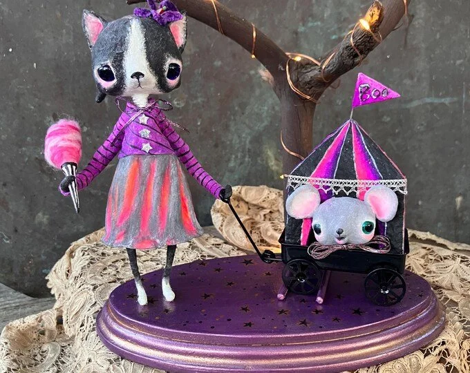 A spun cotton cat and mouse in a toy stroller, set on a purple platform with star patterns. The plush cat is holding a cotton candy, wears a purple dress with a striped top, and is under a small tree with fairy lights.