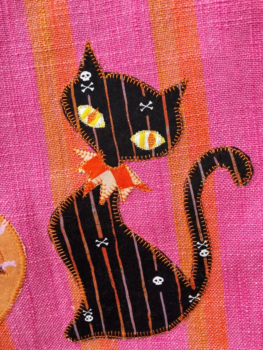 A Halloween-themed fabric patch of a black cat with stitched orange and purple accents, with a background of pink and orange striped tea towel.
