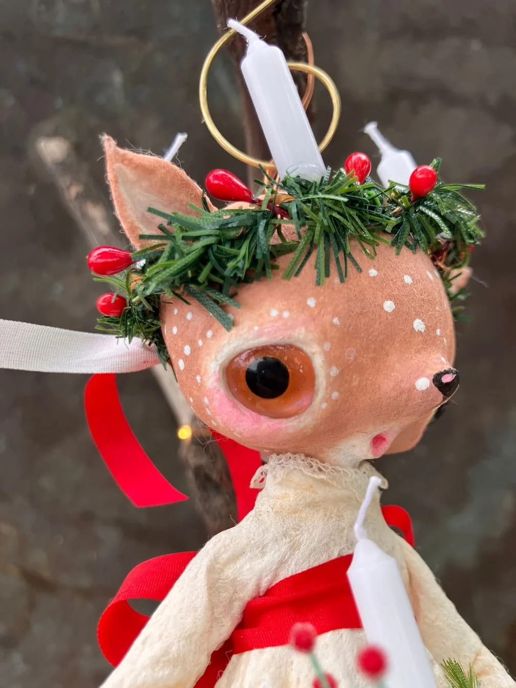 A decorative plush reindeer ornament with large eyes, white polka dots, and a red ribbon, topped with a green holiday wreath and red berries, hanging from a gold hook.