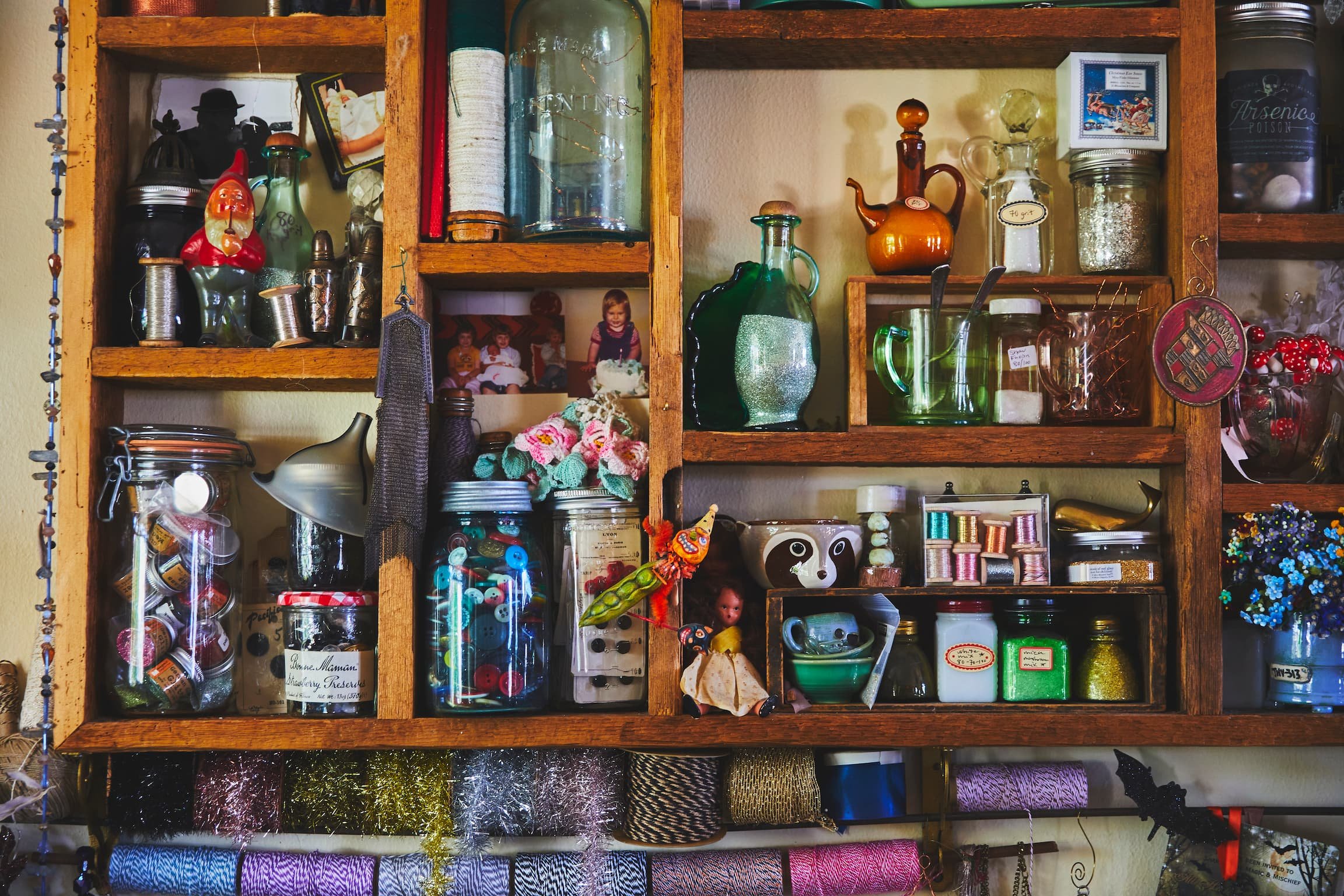 Colorful wooden shelf filled with various jars, bottles, craft supplies, and small decorative objects, including a small Santa figurine, children's photos, and spools of thread.