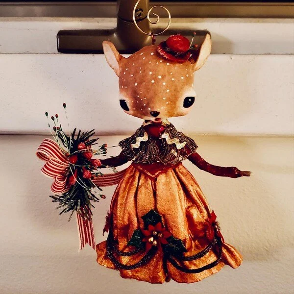 A decorative figurine of a deer dressed in a fancy orange and black dress with Christmas decorations and holding a small bouquet, hanging from a hook on a wall.