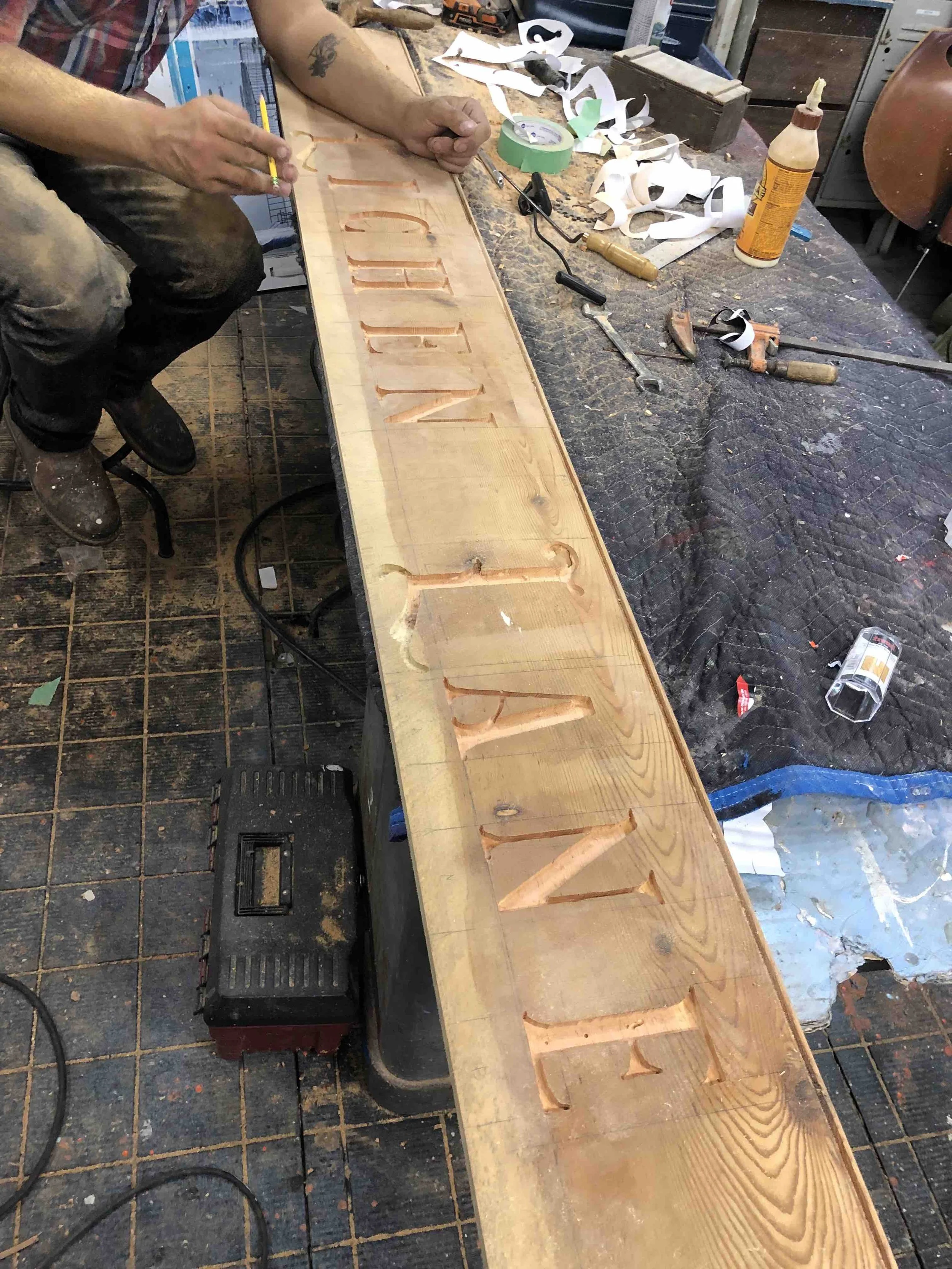 A person is carving letters into a wooden board, with carving tools and supplies scattered on the worktable.
