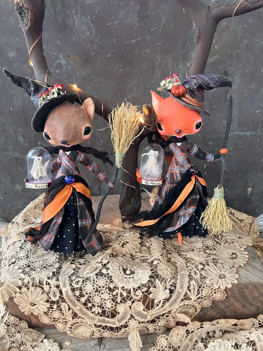 Folk art Halloween decorations featuring two anthropomorphic animal figures dressed as witches, holding miniature broomsticks, with mushroom and owl motifs, placed on a lace cloth.