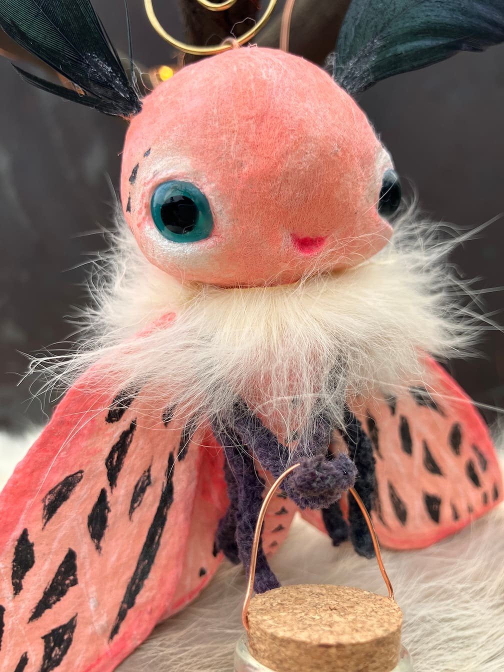 A plush toy with a large head, blue eyes, and a pink face, dressed as a butterfly with pink, black, and white wings and black antennae with golden hoops, holding a small cork with a wire handle.