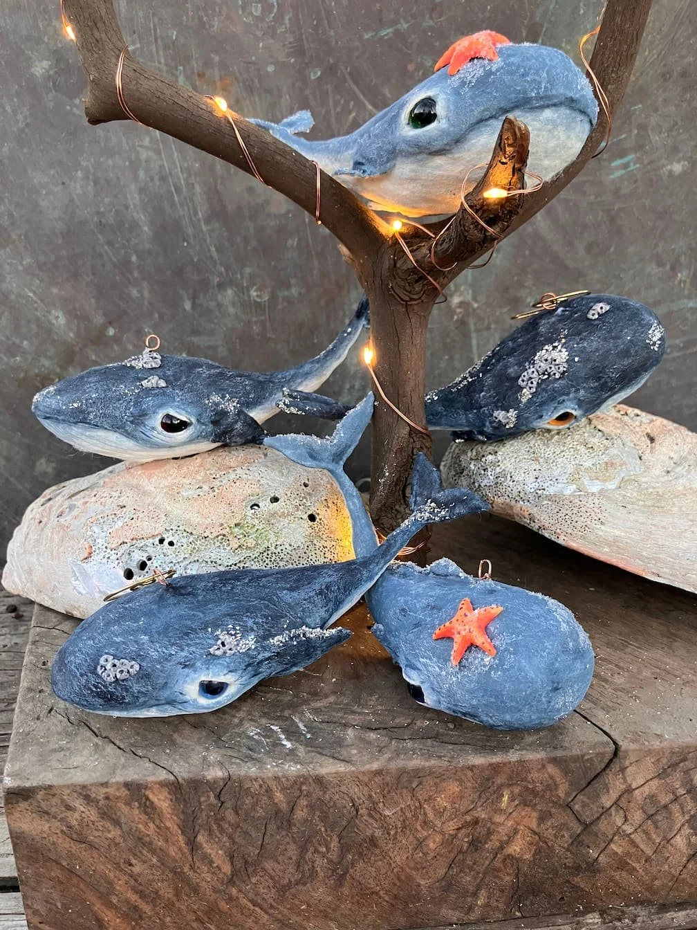 Decorative display of spun cotton whale ornaments holding starfish. Below are rocks and small tree branches with string lights, arranged on a wooden surface.