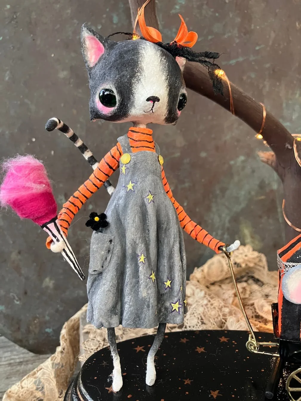 A cute cat with a painted face, pink cheeks, and big black eyes, dressed in an orange and black striped long-sleeve shirt and a gray dress decorated with yellow stars. The doll has a pink and black striped tail, a black flower on its dress, and is ho