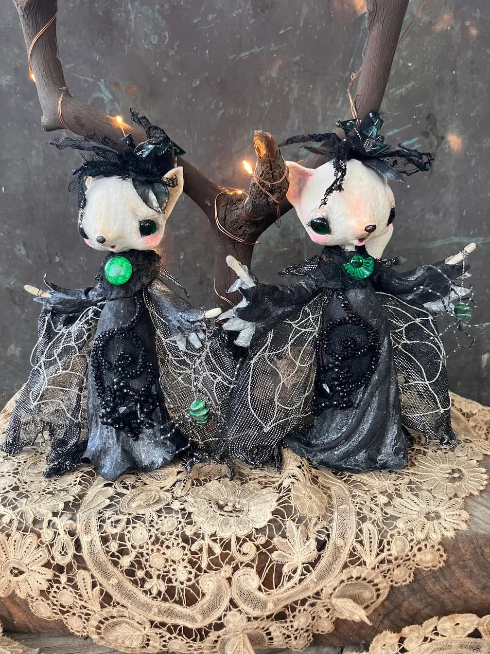 Two fairy mouse spun cotton ornaments with black lace and tulle dresses, green gemstone necklaces, and black ribbon bows on their heads, hanging from a twisted branch with fairy lights, placed on a lace doily.