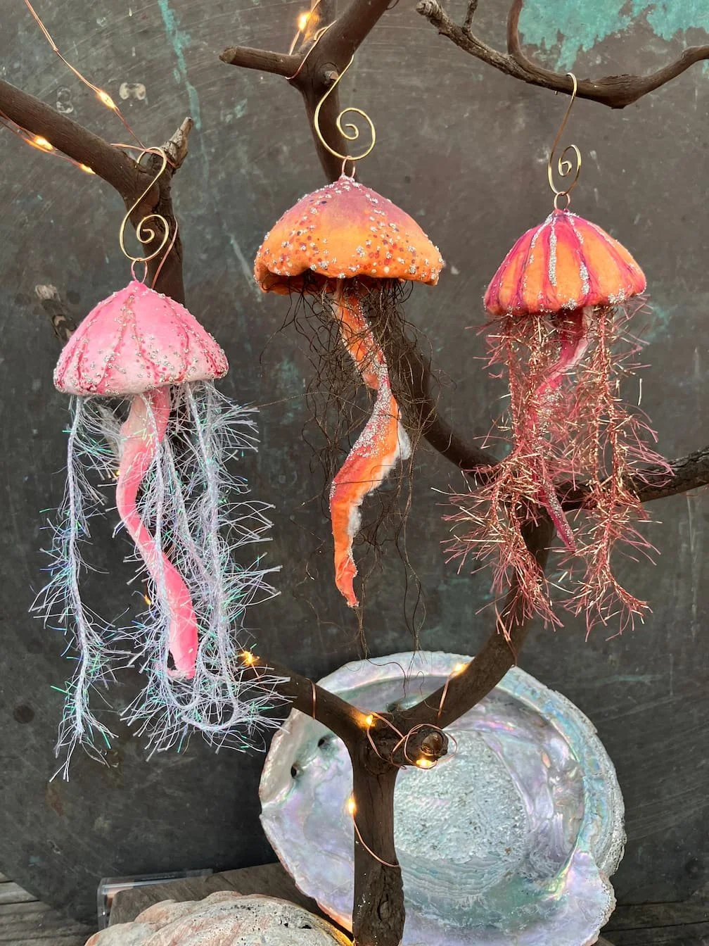 Decorative hanging jellyfish ornaments made of spun cotton, sparkling ribbons, wire, and beads, hanging from a branch with wire lights, against a dark background.