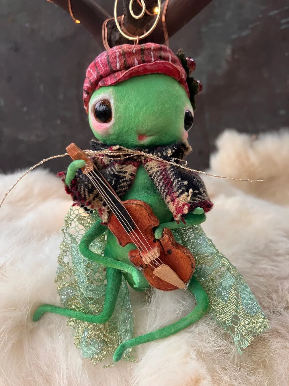 A green puppet with large black eyes, dressed in a red plaid hat, a black and red scarf, and lace wings, holding a small wooden violin.