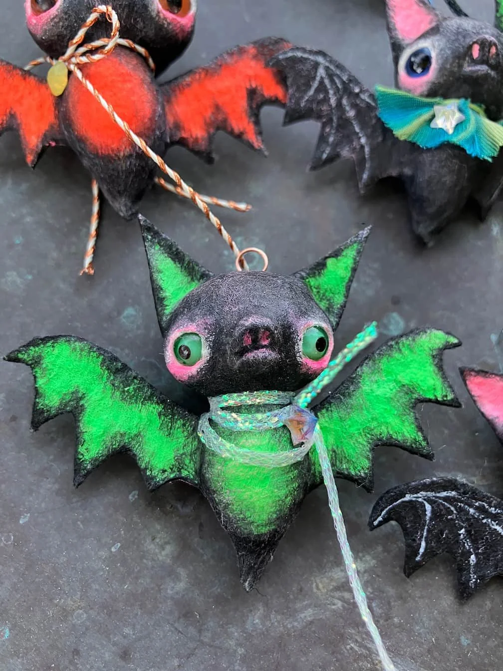 Colorful plush bat decorations with big eyes, green and black wings, and pink cheeks on a dark surface.