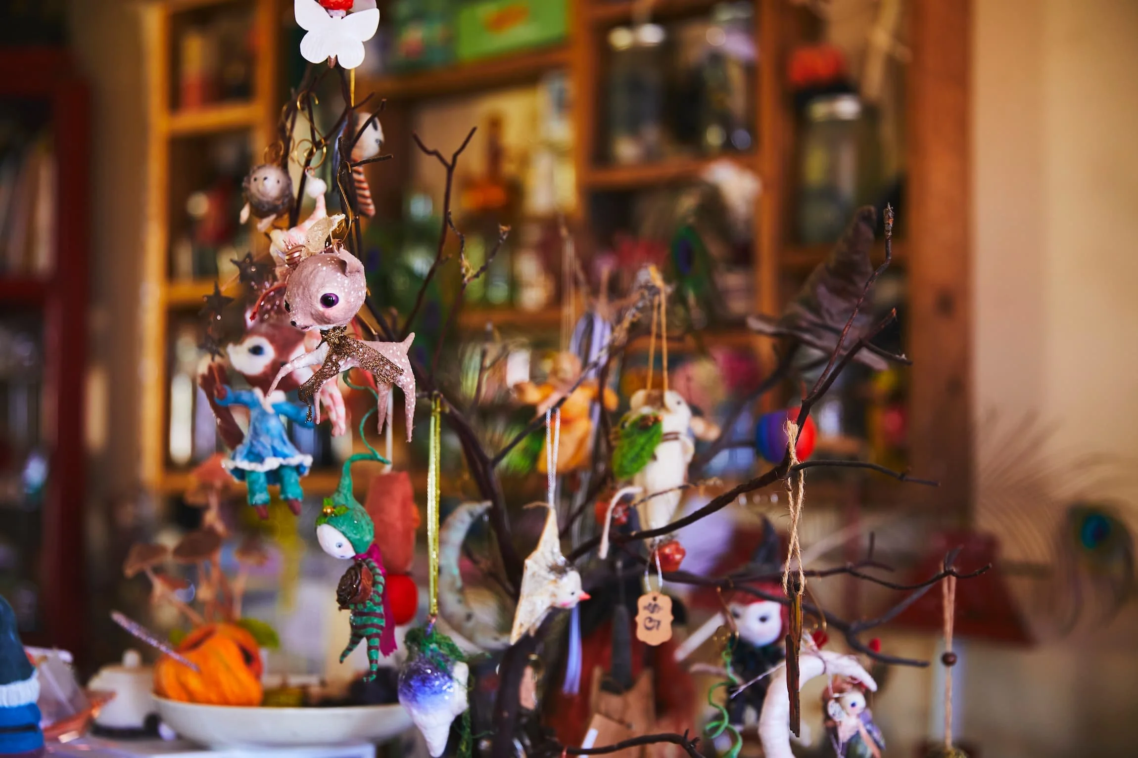 Colorful handmade cherub and fairy ornaments hanging from bare tree branches on display, with shelves filled with various items in the background.