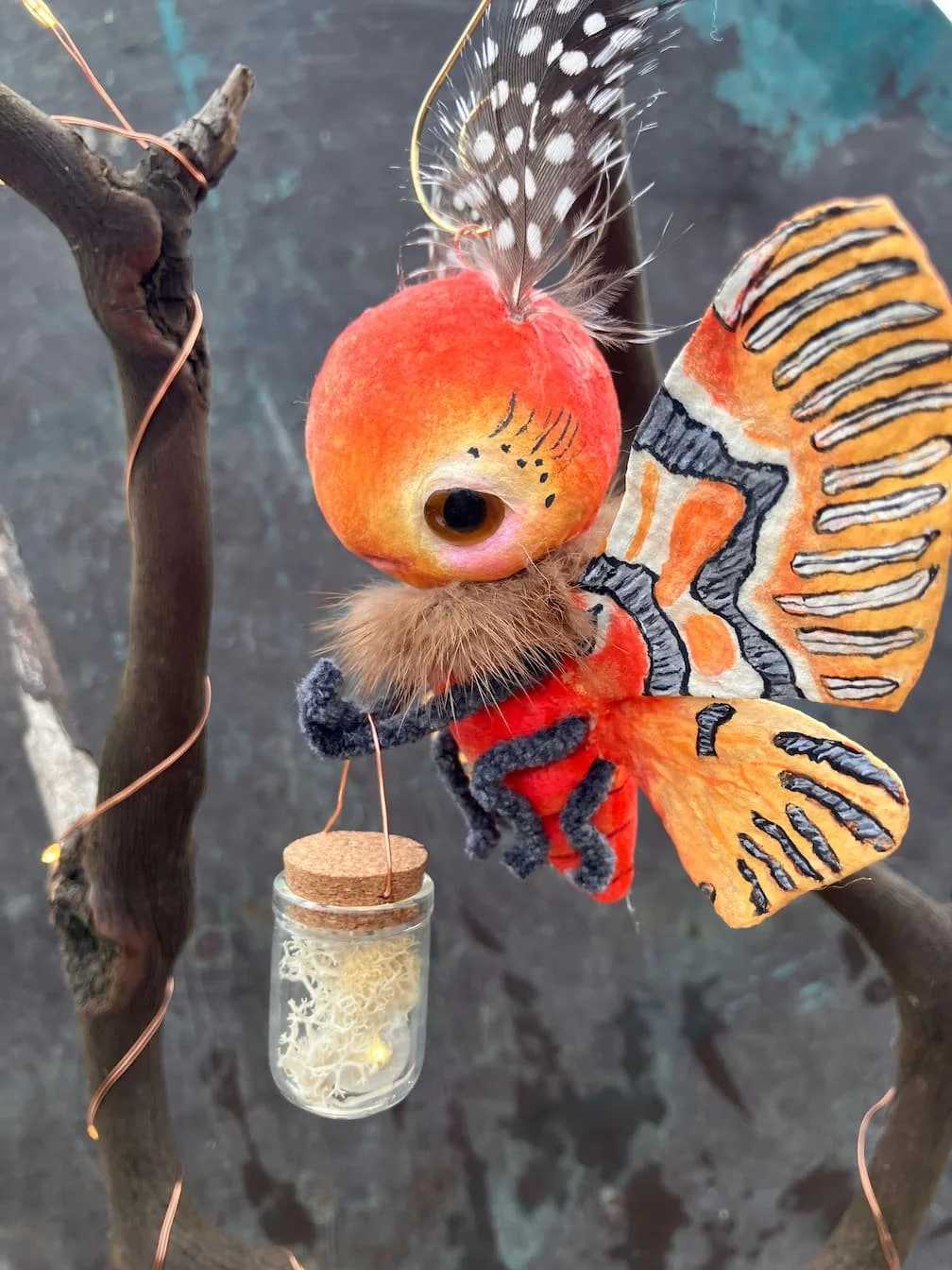 A handcrafted art doll resembling a butterfly with a peach face, feathered antennae, and detailed orange, black, and white patterned wings, perched on a tree branch with a small glass jar hanging below.