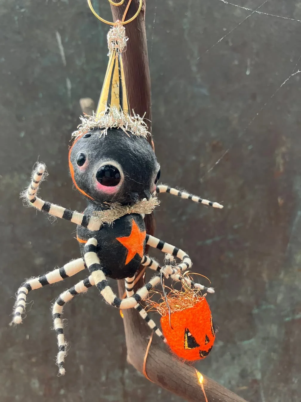 A black spider with orange star on its body and striped legs, wearing a tiny gold crown, hanging from a branch with Halloween-themed decorations.