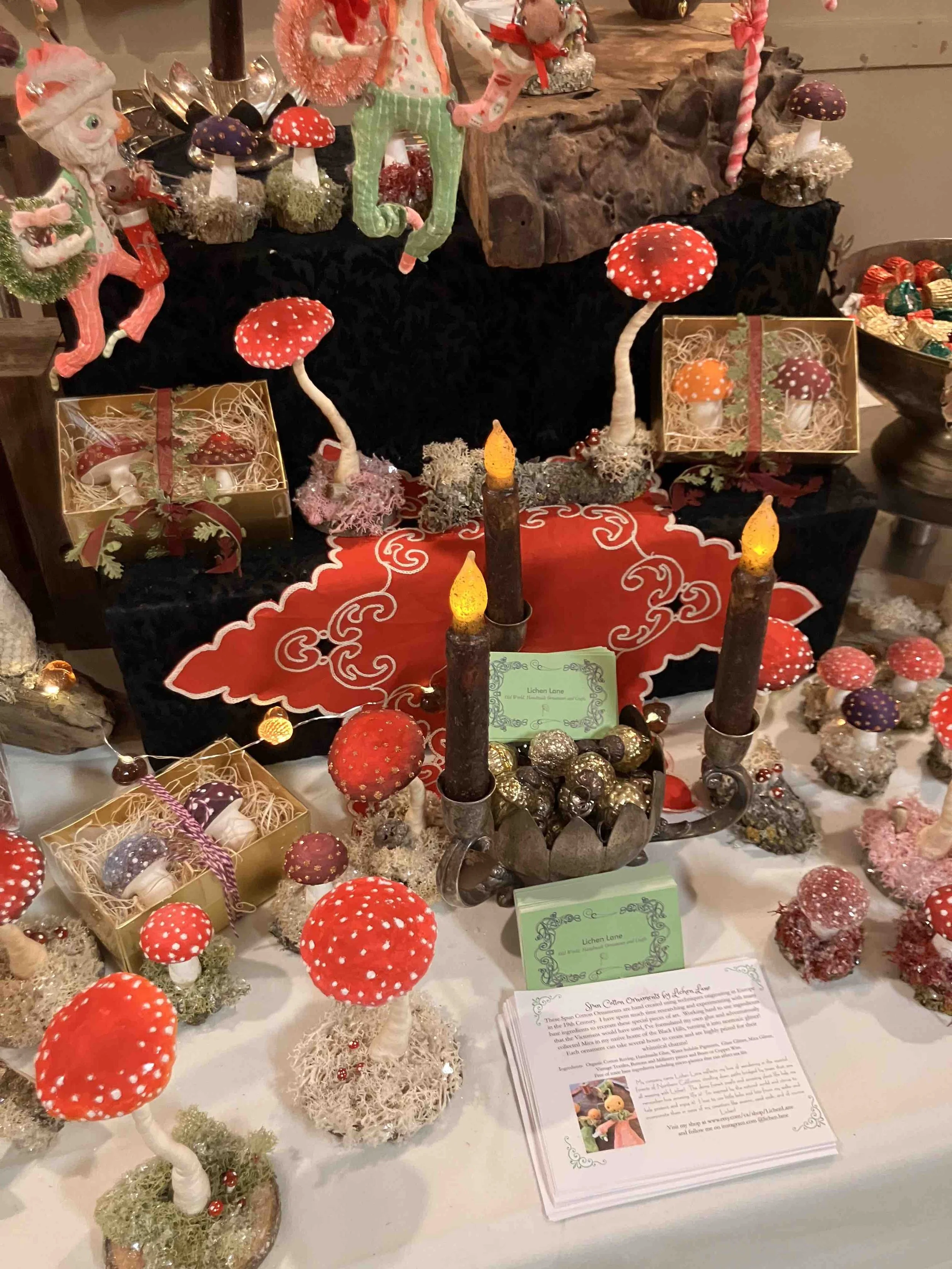 Display of handmade mushroom ornaments, fairy figurines, and decorative items on a table, with informational pamphlets nearby.
