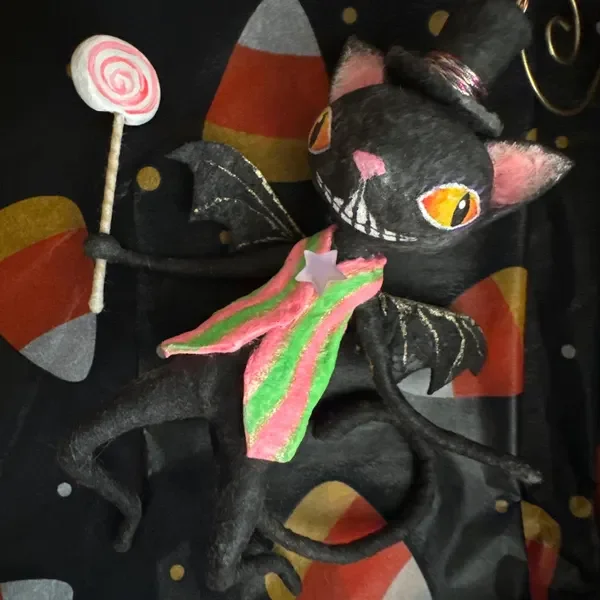A black spun cotton bat with orange eyes, pink nose, and stitched mouth, wearing a rainbow-colored scarf and a small pink star on its chest, holding a candy-shaped lollipop, against a black fabric background with colorful patterns.