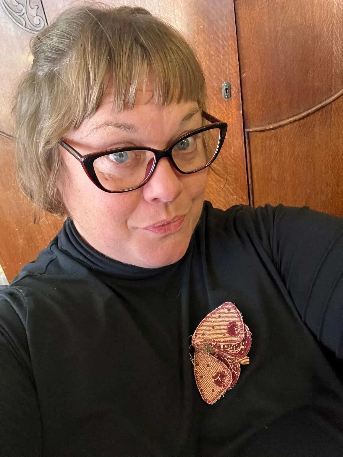A woman with short light brown hair and glasses taking a selfie in front of a wooden background, wearing a black top with a decorative bow.