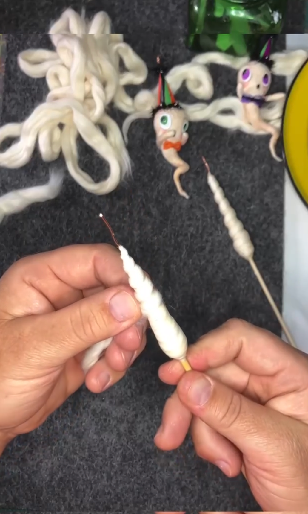 Close-up of hands twisting white yarn to create a coiled, handmade spun cotton object with a cone-shaped face and a pointy hat. The background shows more yarn and some completed doll features, with various decorative eyes and accessories.