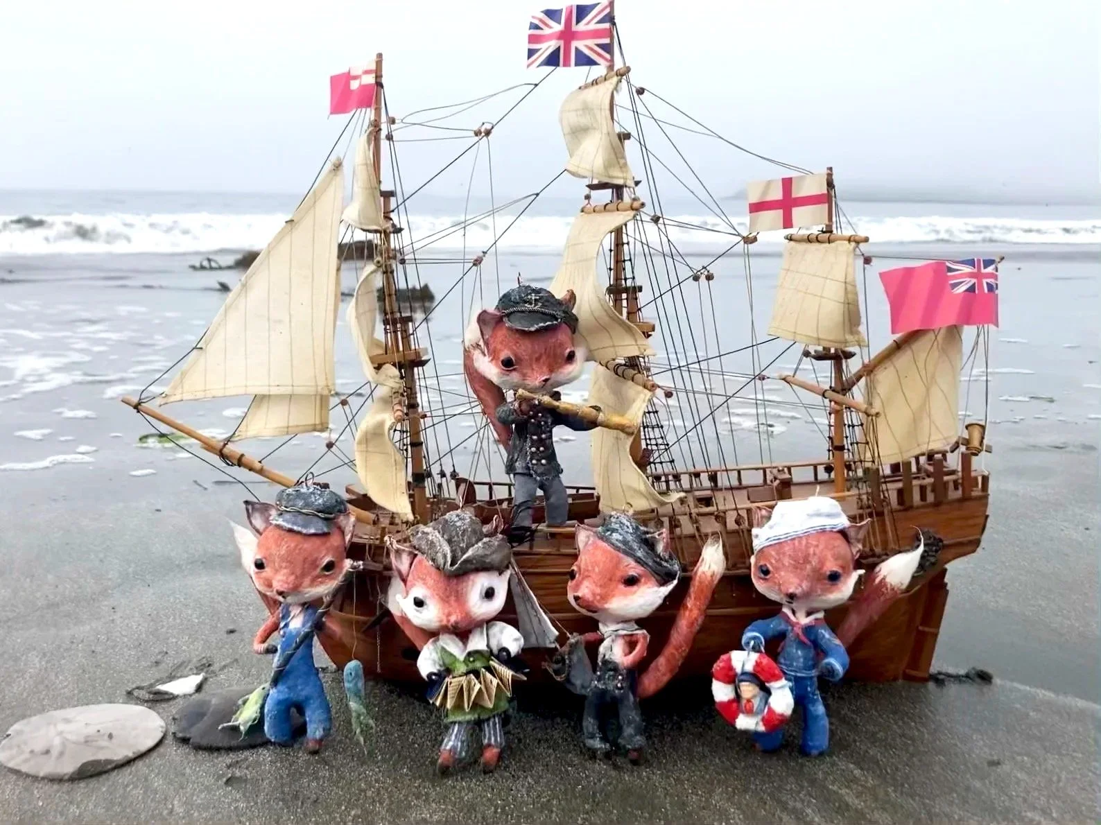 A toy ship with spun cotton ornament animal characters dressed as sailors on a beach, with a background of ocean waves.