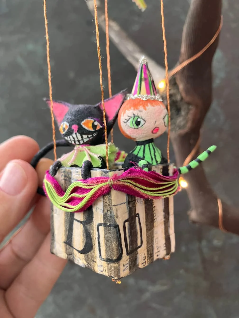 Miniature puppet theater with hand puppets of a black cat with orange eyes and a winking face, and a girl with green eyes, red lips, and a pink party hat, sitting inside a decorated box with colorful fabric and string strings.