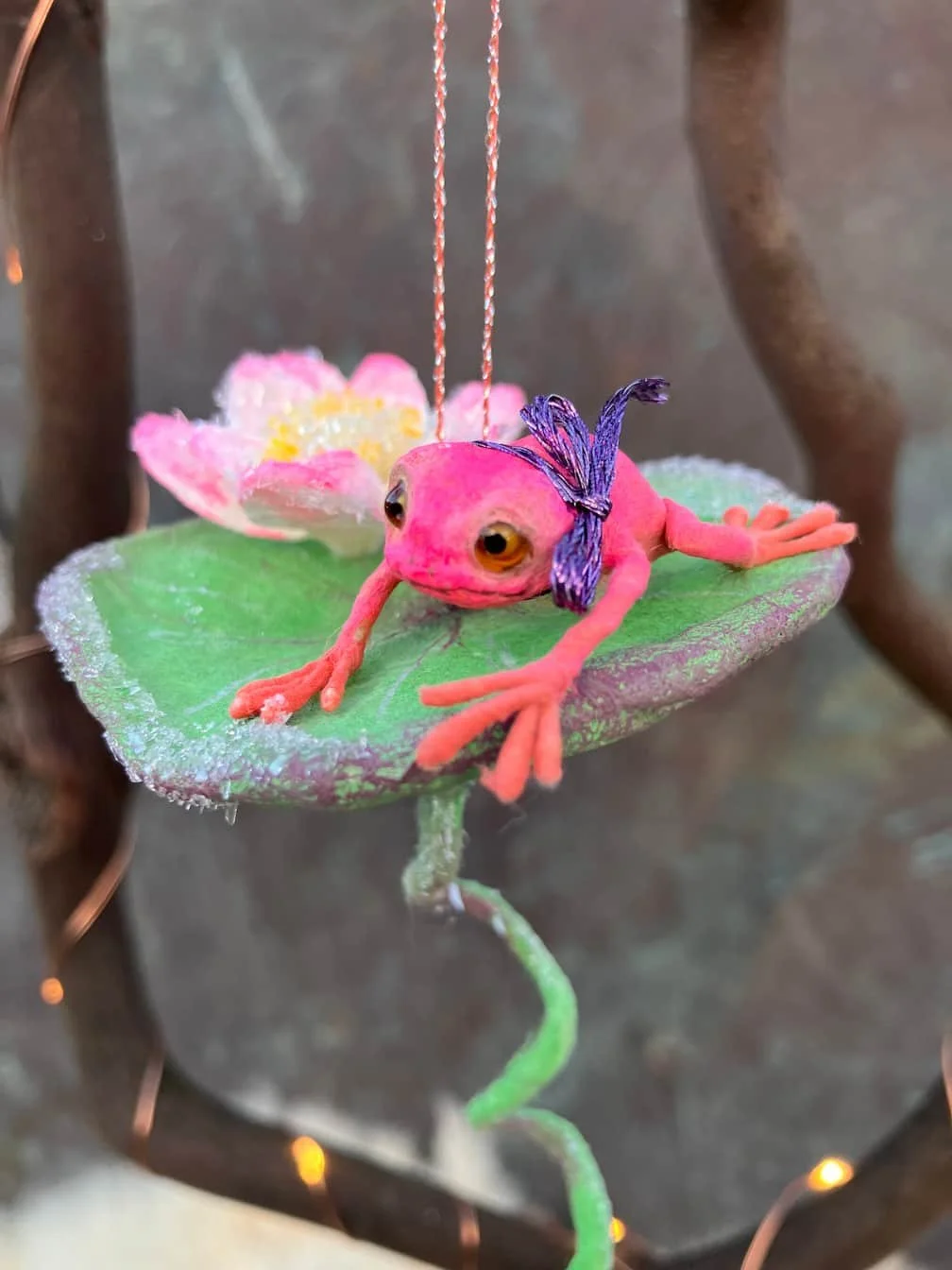 A small decorative ornament resembling a frog with large eyes, lying on a green leaf with pink and white flowers in the background, hanging from a pink and white string.
