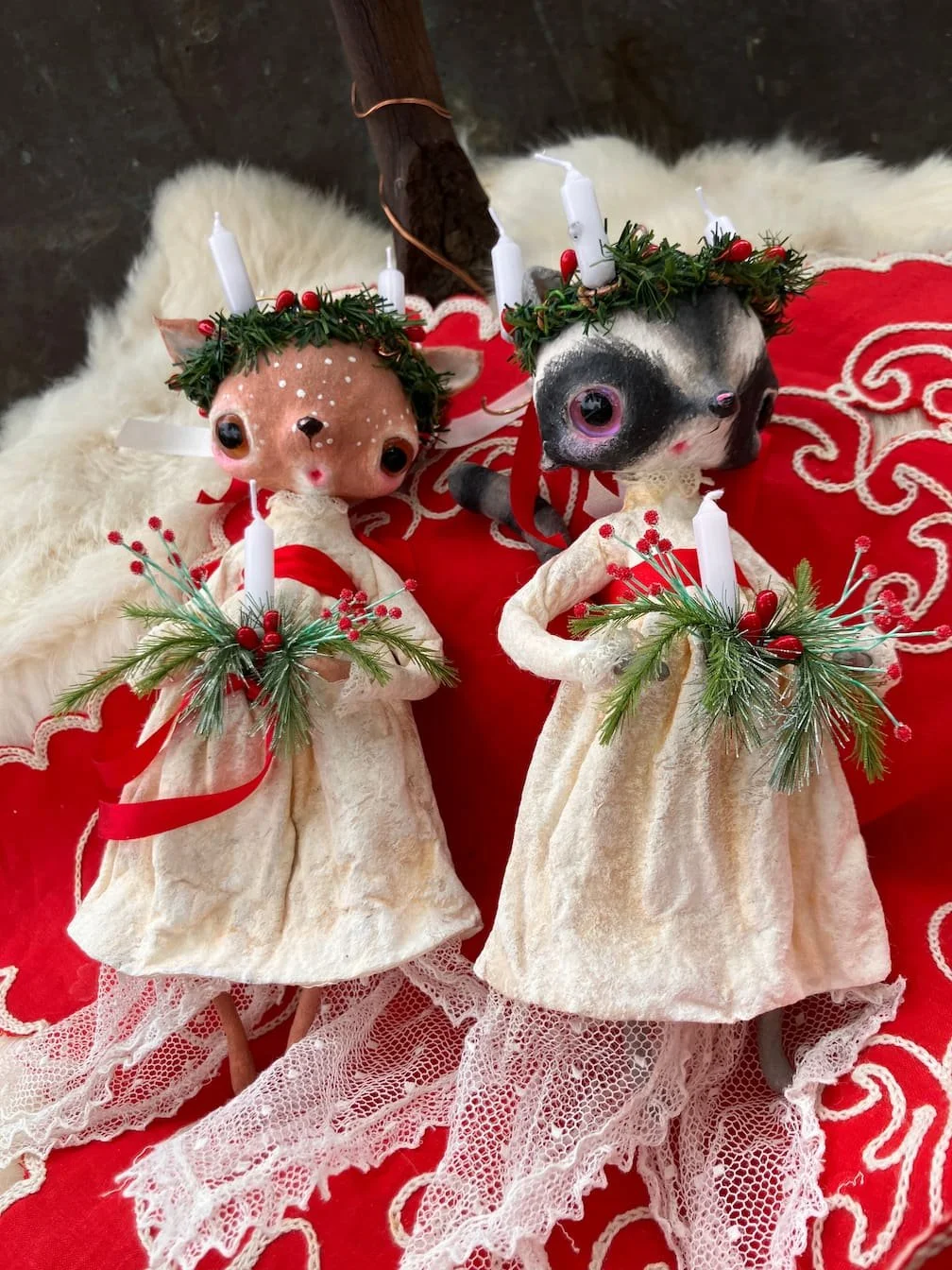 Decorative dog and cat ornaments dressed in white dresses with Christmas greenery and candles, placed on a red embroidered cloth.