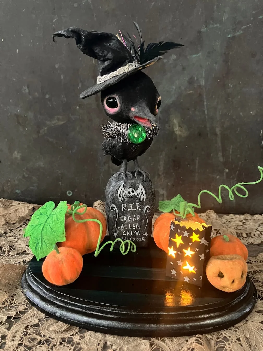 A Halloween-themed display with black bird figurine wearing a witch's hat, decorated with a green jewel necklace, standing on a tombstone marked 'R.I.P. Edgar Allen Crow 2023.' Artificia, all arranged on a black oval base with lace fabric underneath.