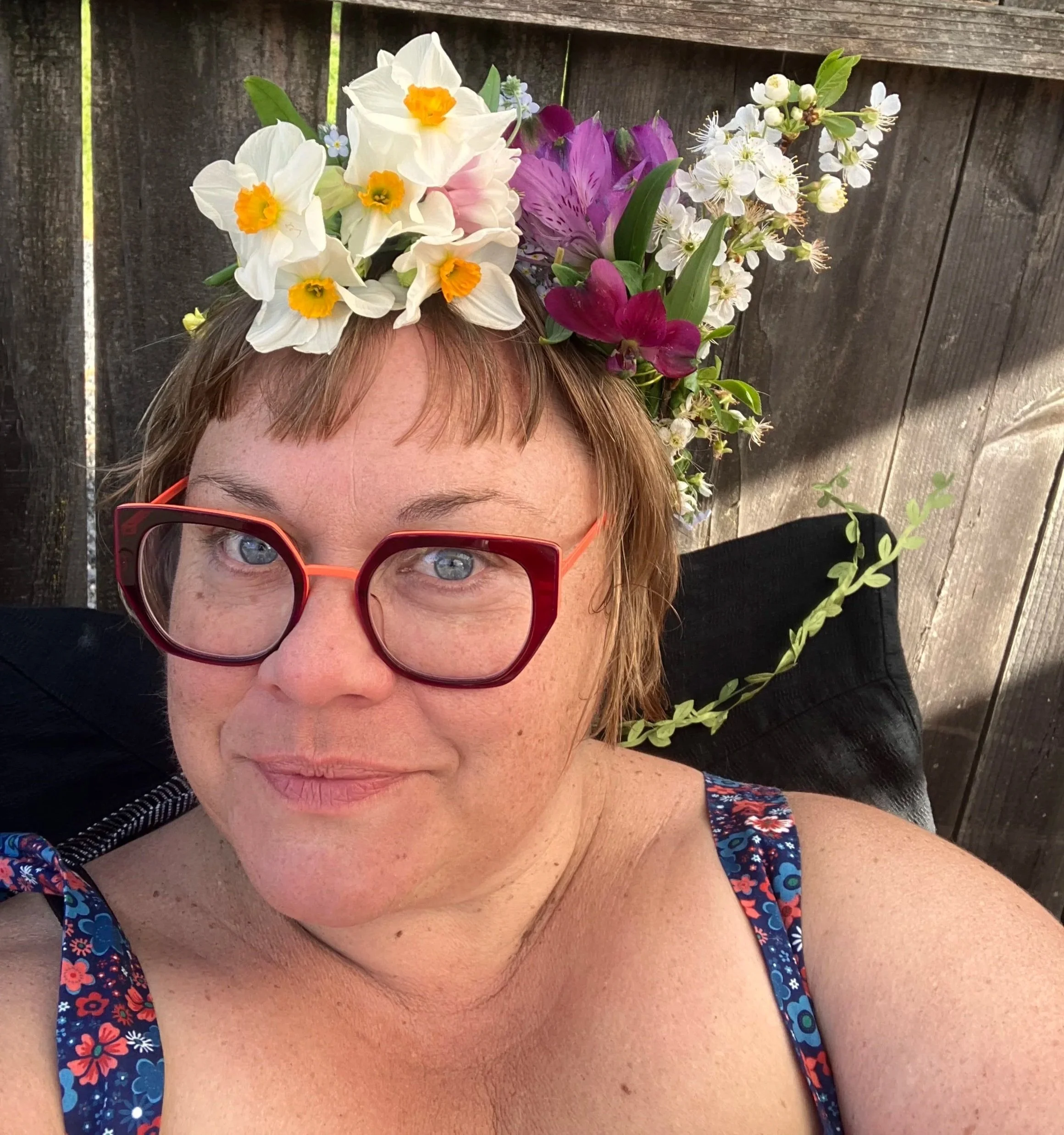 A woman wearing red glasses and a floral dress is lying down outdoors with her head resting against a wooden fence. She is wearing a crown made of blooming flowers, including white daffodils, purple orchids, and small white blossoms, with some greenery.