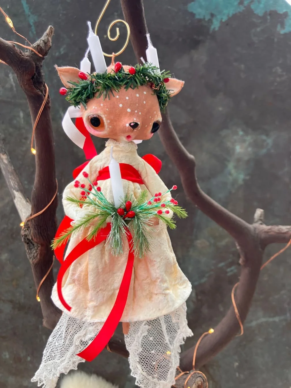 A cute deer figurine dressed in a cream dress with lace trim, decorated with Christmas wreaths, red ribbons, and holly berries, hanging on a tree branch.