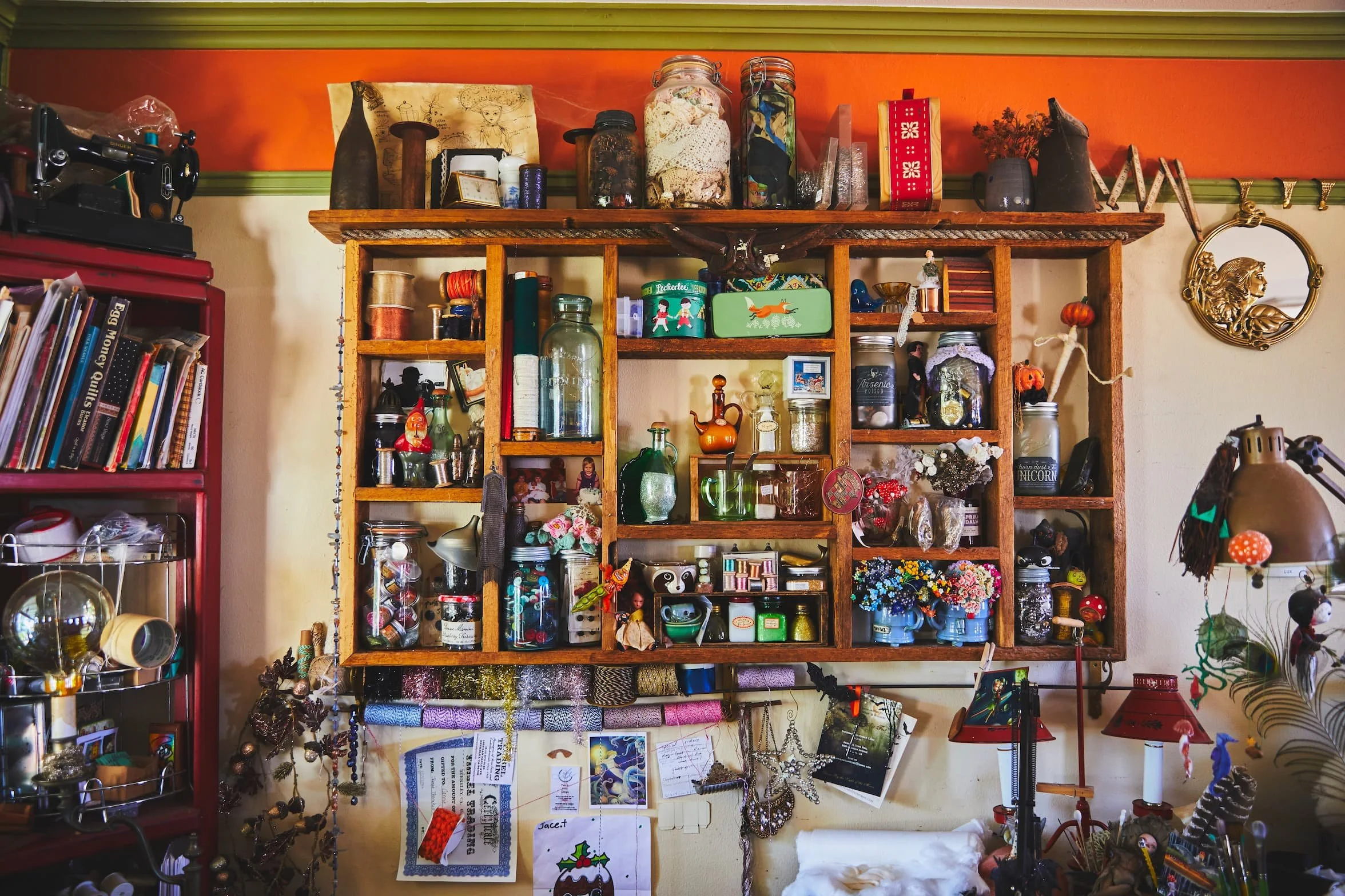 A wooden wall shelf with multiple compartments holding jars, decorations, small figurines, and various knickknacks. To the right, a decorative lion face wall mirror is visible. On the left, part of a red bookshelf with books and a metal rack with jars can be seen. The background wall is painted in cream with a horizontal colorful border.