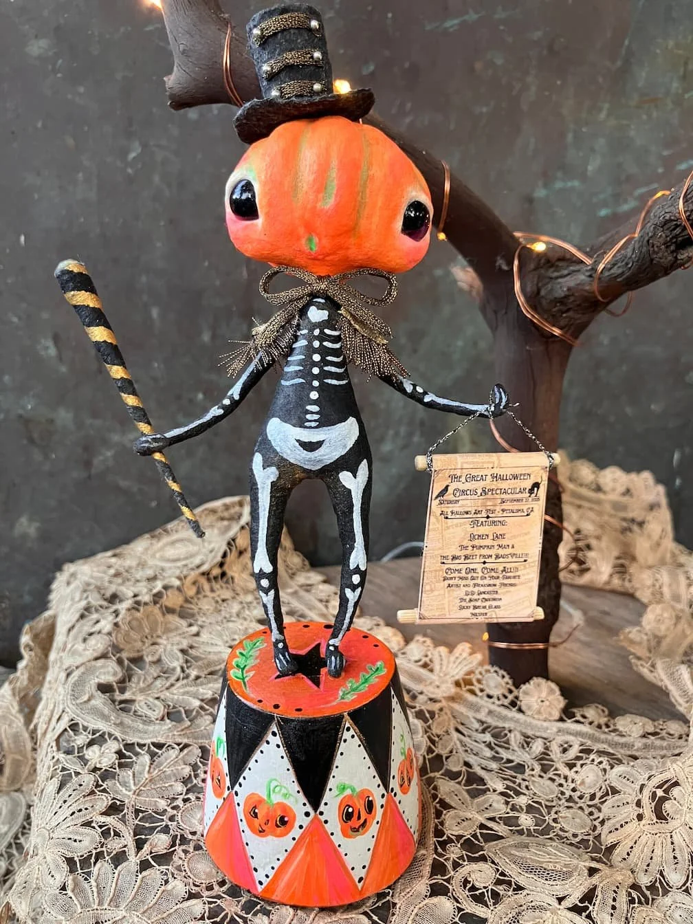 Decorative Halloween figurine of a cat with a pumpkin head, wearing a black and white skeleton costume, holding a striped tail and a sign about a Halloween event, standing beside a small Halloween-themed carousel base with pumpkins. In the background