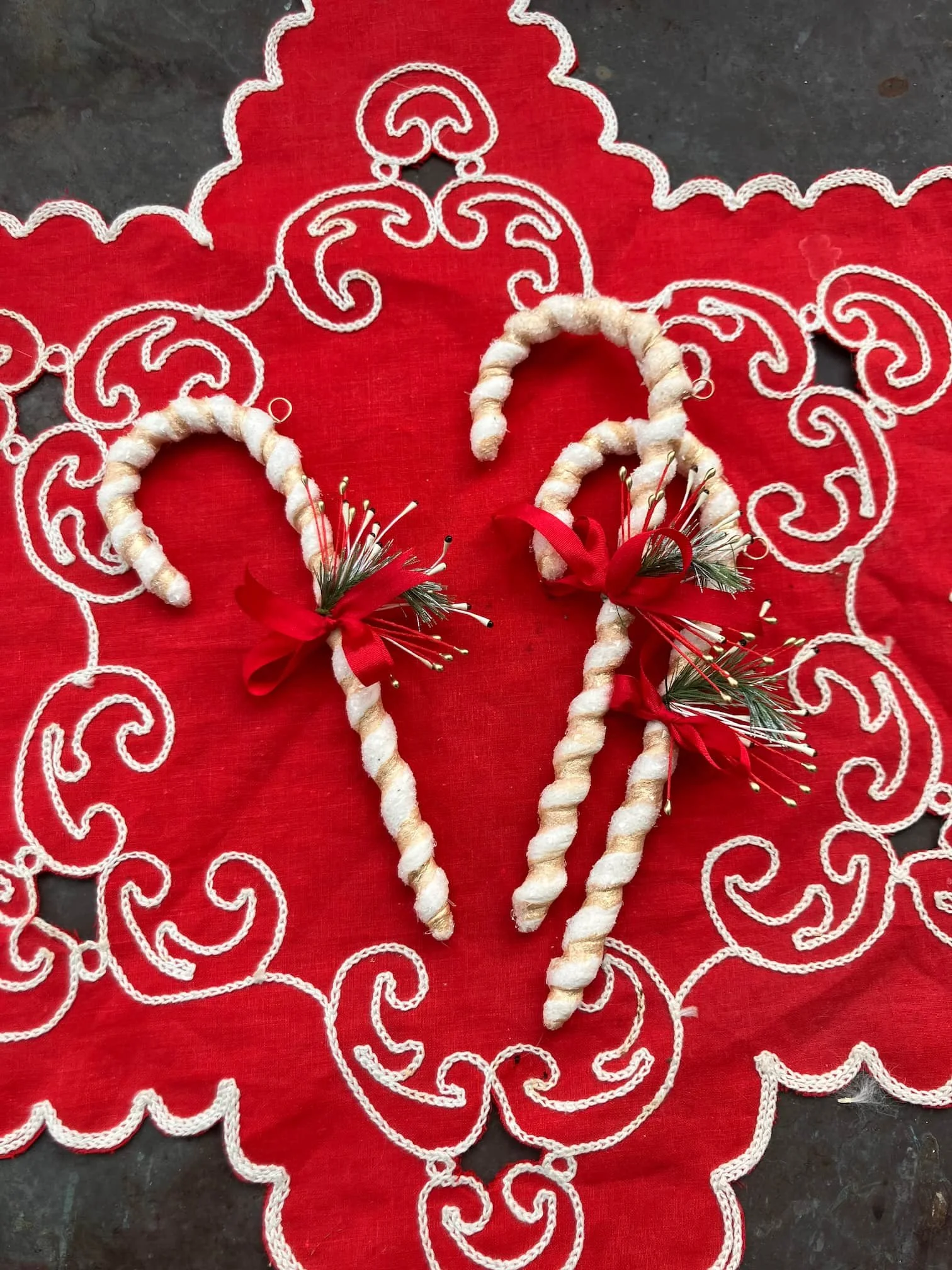 Two decorative angel-shaped holiday ornaments wrapped in white thread, decorated with small artificial pine branches, red ribbons, and small white stars, placed on a red embroidered tablecloth.