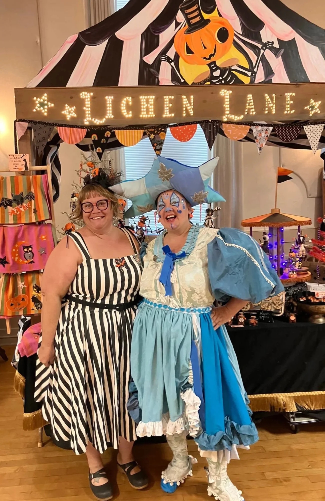 Two women are dressed in Halloween costumes, standing in front of a decorated booth One woman is wearing a black and white striped dress with glasses, and the other is dressed as a large blue and white fairy or princess with elaborate face makeup and