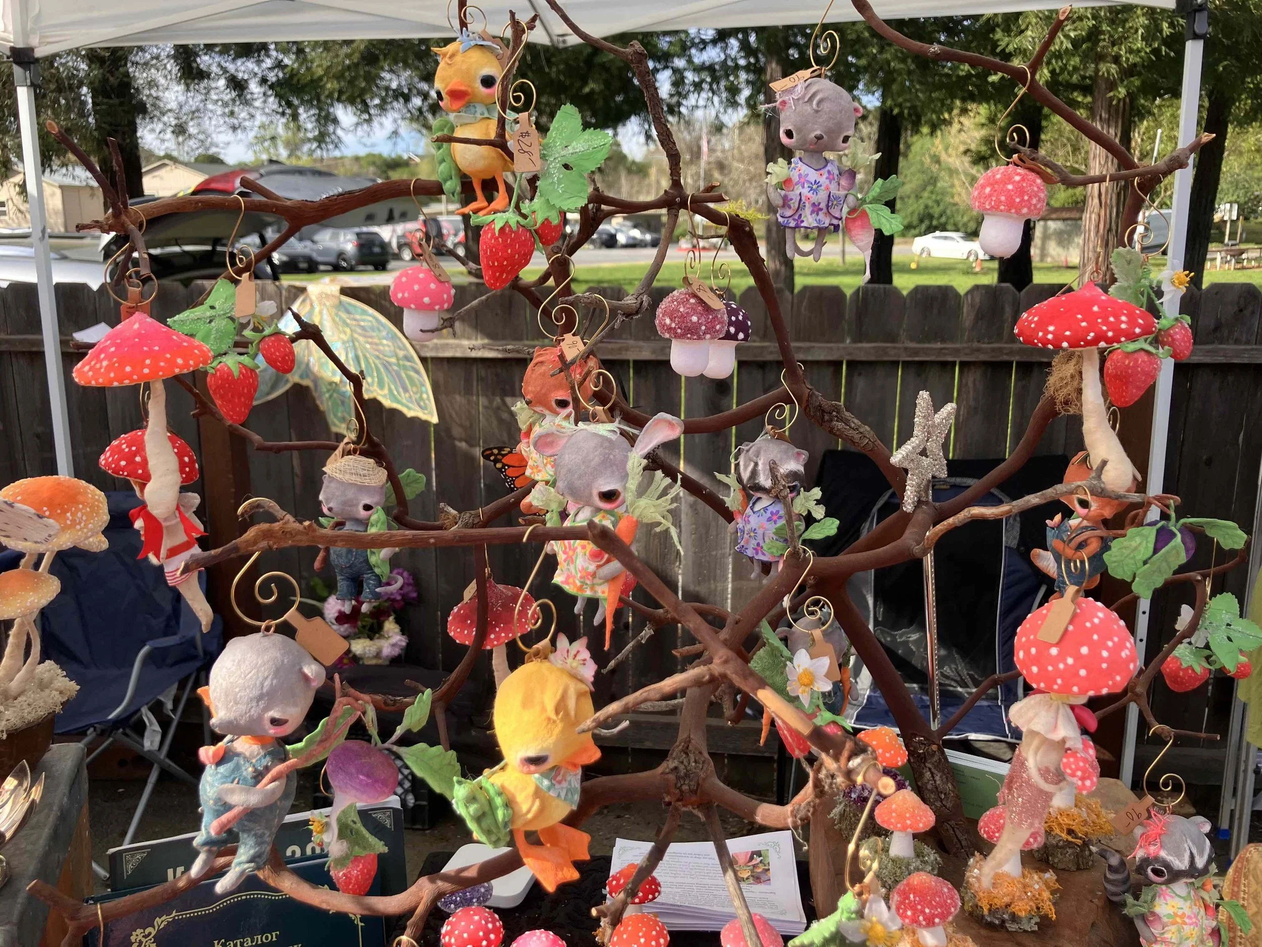 A display of whimsical handmade animal and mushroom ornaments hanging from a tree branch, including a bird, koala, mouse, fox, and various mushrooms, at an outdoor market or craft fair.