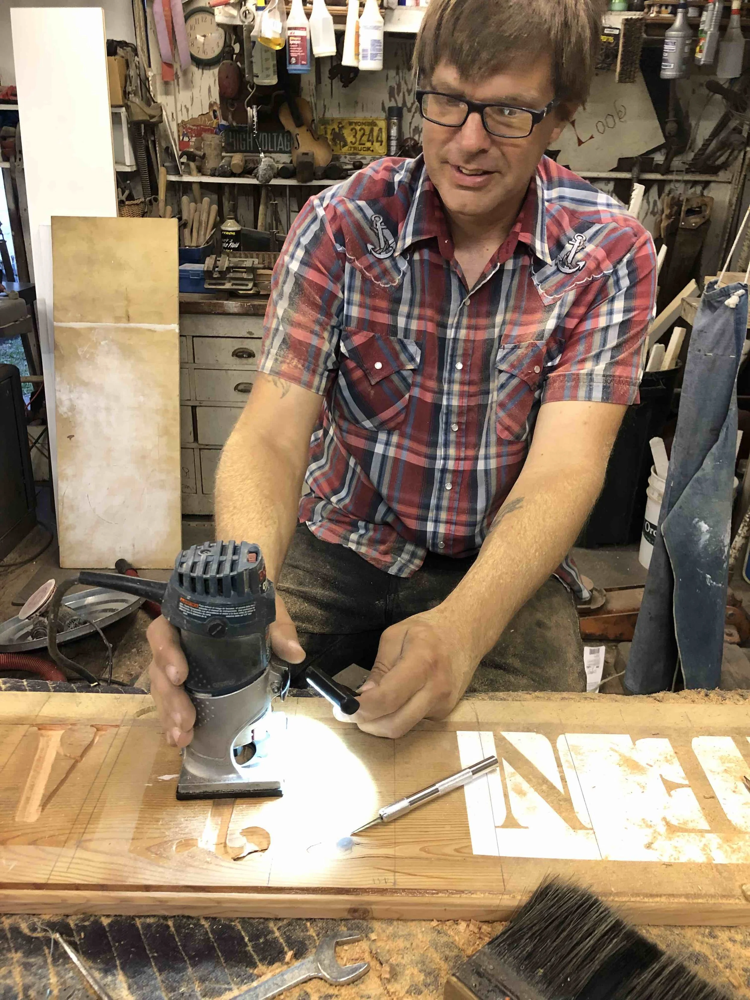 A man working in a woodworking shop, using a router on a piece of wood, surrounded by tools and supplies.
