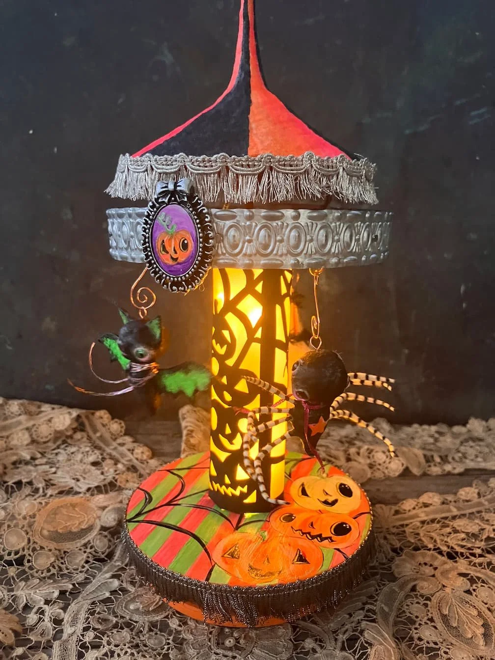 Halloween-themed decorative lantern sculpture with pumpkins and spider decorations, featuring a striped base, a glowing central column, and a small top hat with a pumpkin face