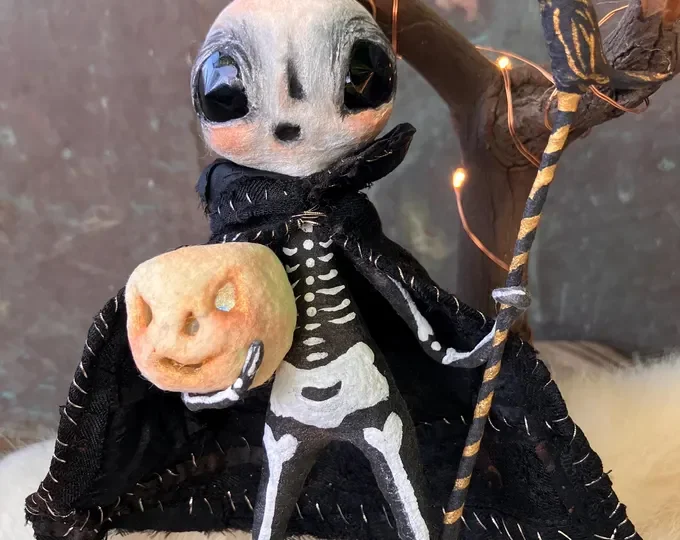 Cute skeleton Halloween figure holding a smiling apple with a carved face, decorated with black cloak and fairy lights.