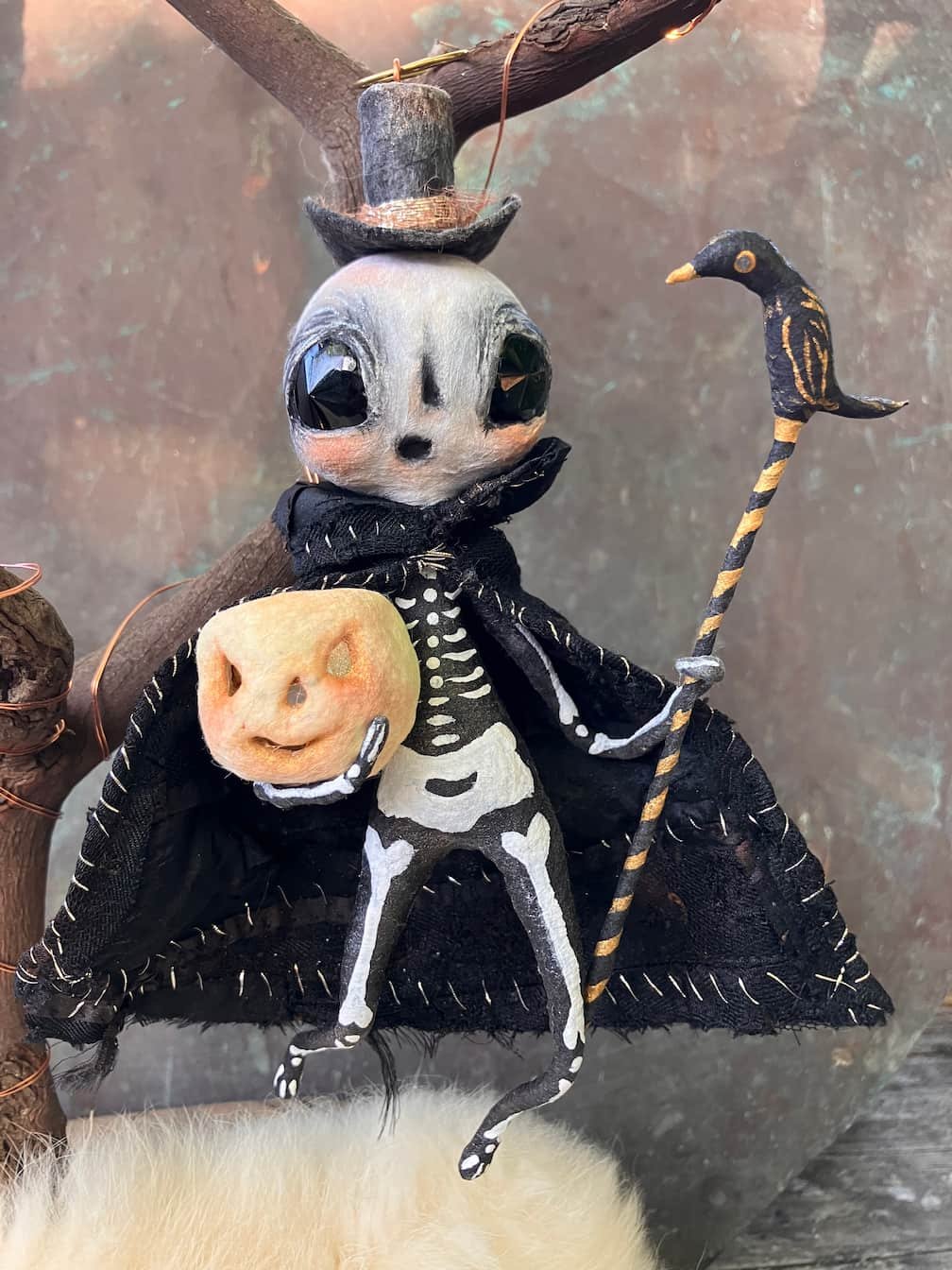 A Halloween-themed doll featuring a skeleton body wearing a black cloak, with a skull head that has big, dark eyes and a small nose. It holds a small apple-shaped object with a carved face in one hand and a black and gold striped staff with a black b