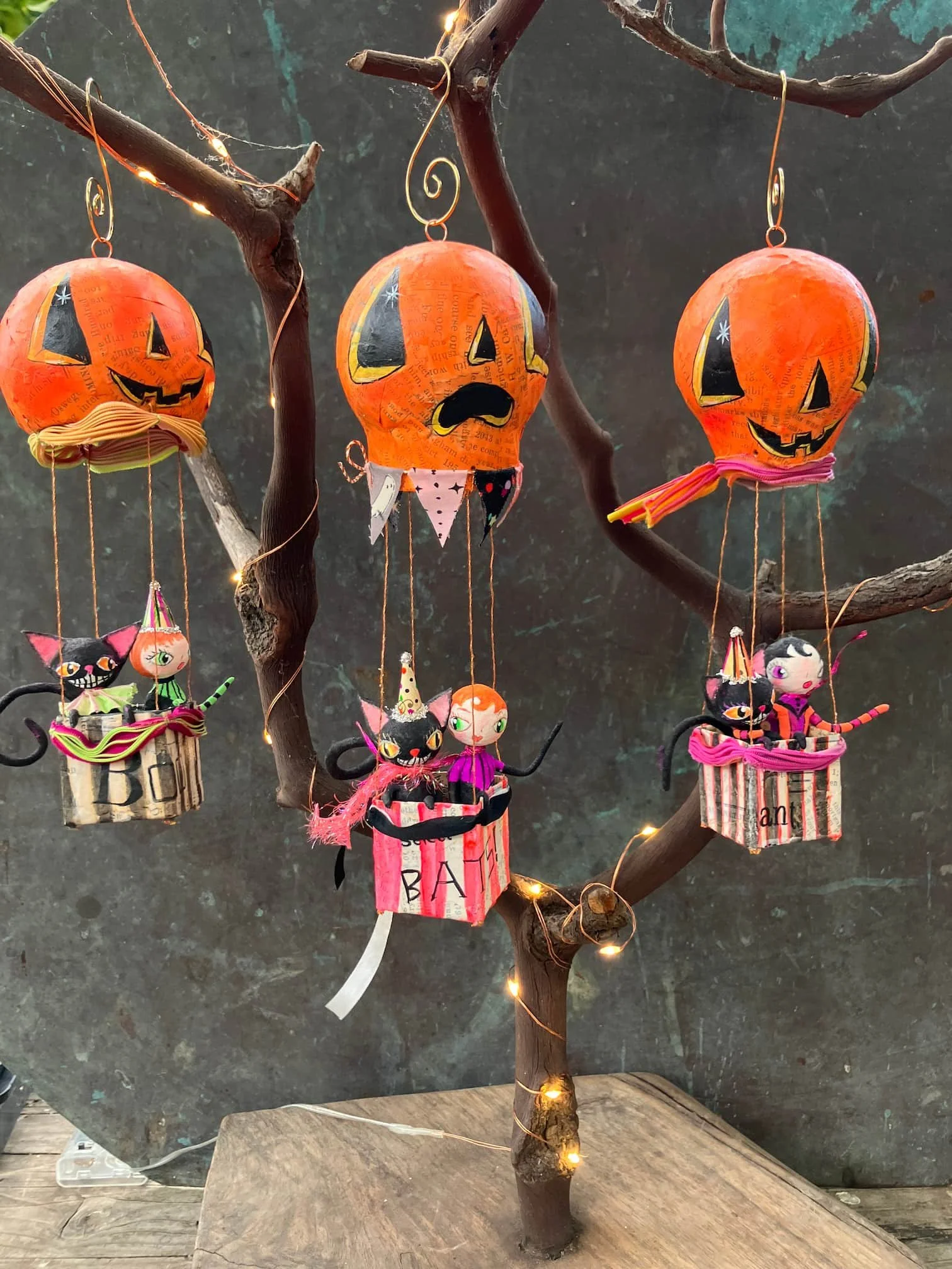 A decorative display of Halloween-themed lanterns hanging from a small tree with fairy lights. The lanterns are orange and feature various faces of Jack-o'-lanterns and other spooky characters, some with colorful details and accessories. The backgrou